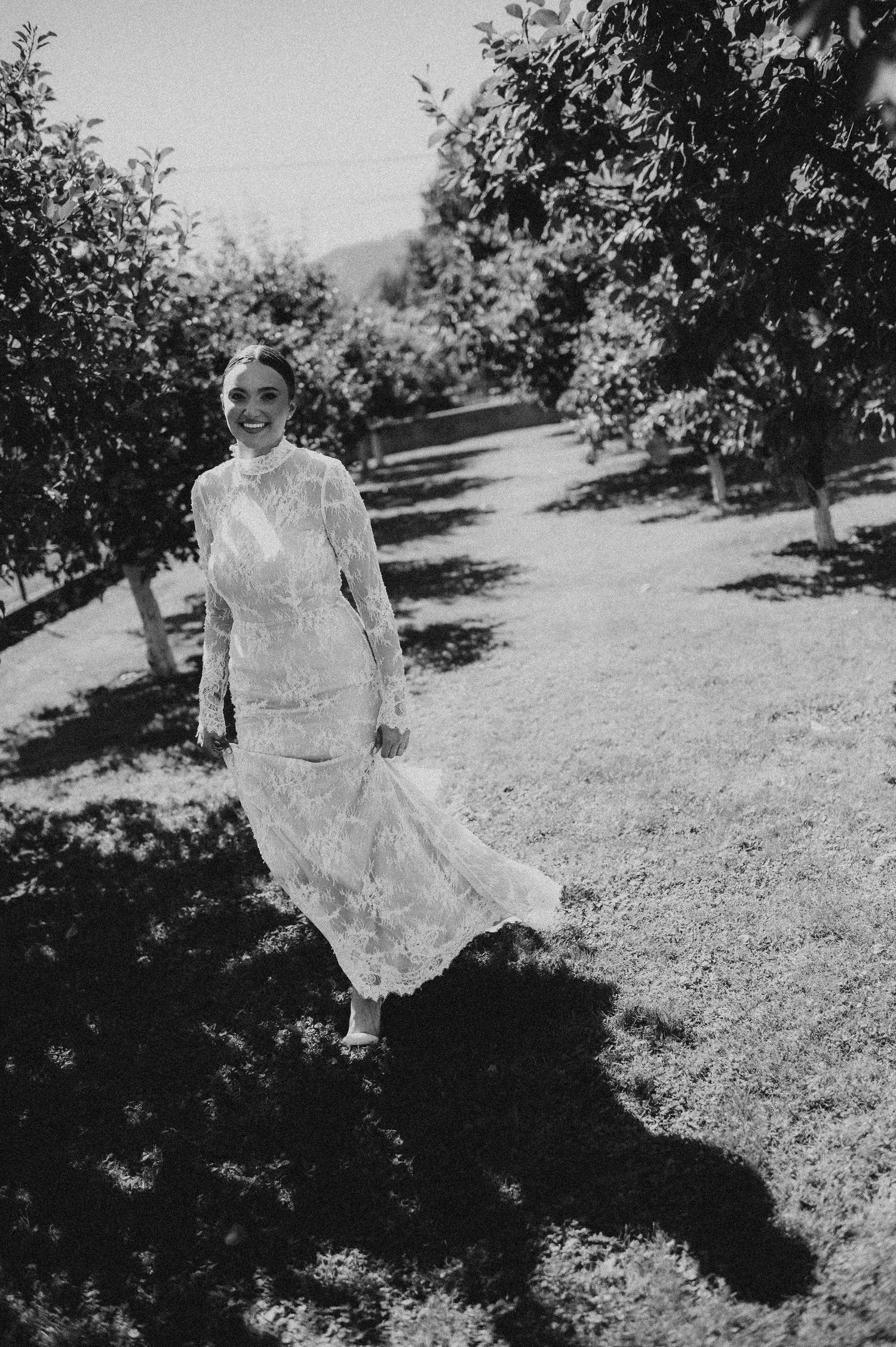 Bride in a long-sleeve lace wedding gown walking through a sunlit orchard path in black and white.