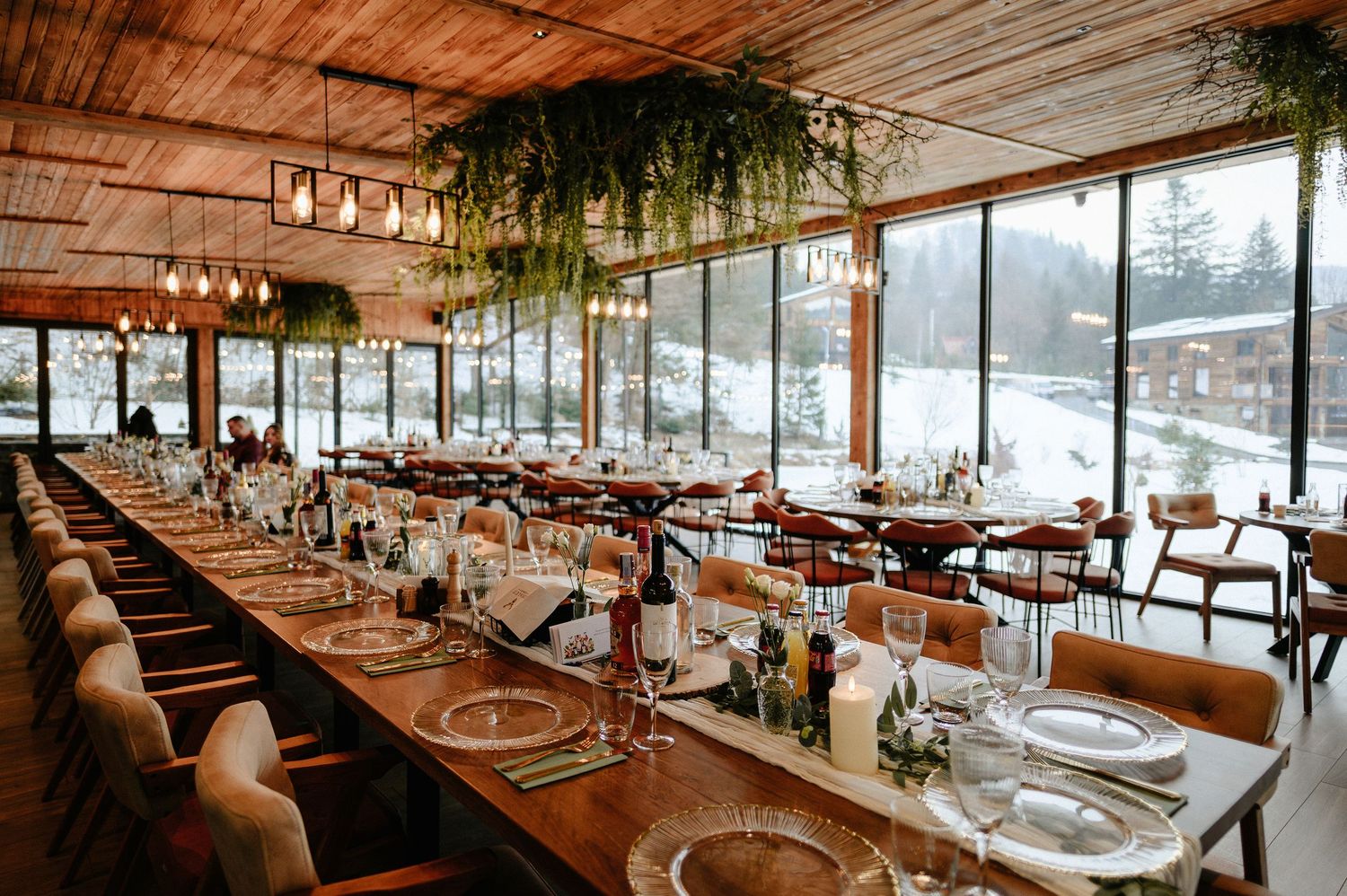 Elegant long banquet table set for a winter wedding reception inside a rustic wooden venue with large windows.