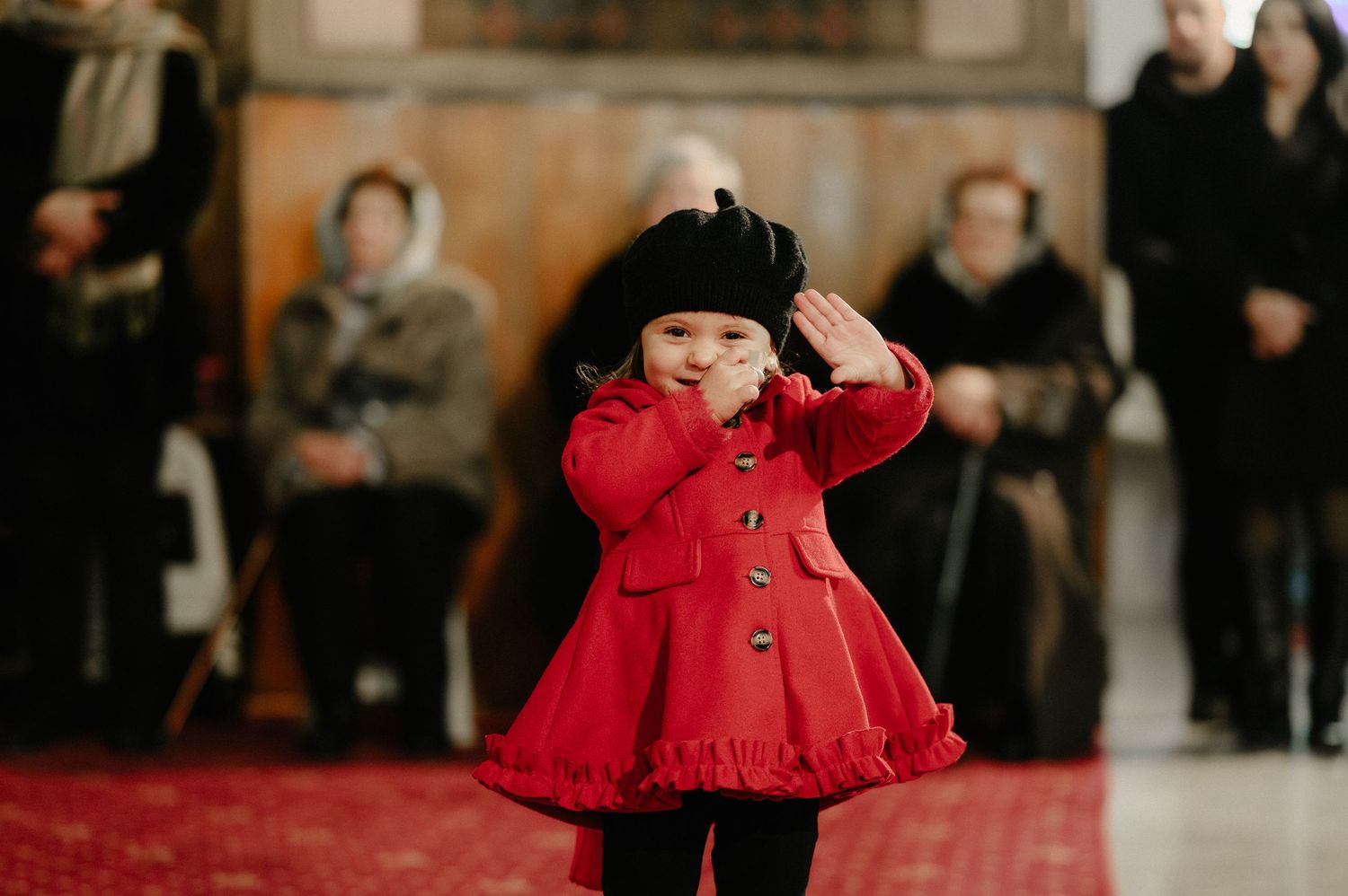 Adorable toddler girl in red coat and black beret walks down a church aisle during a ceremony.