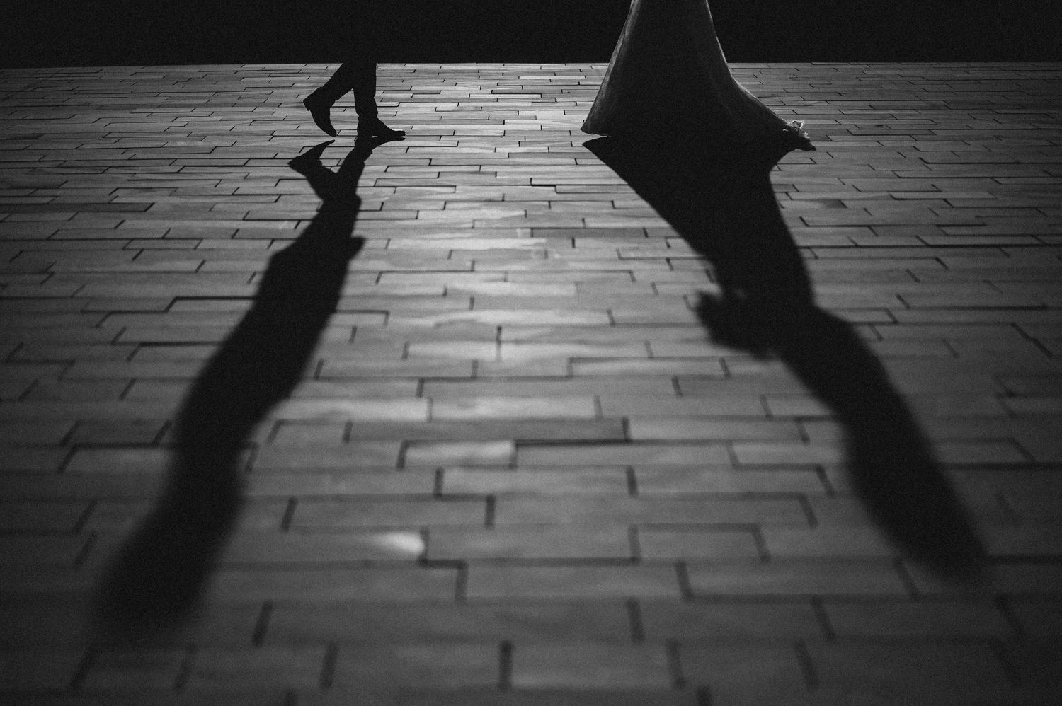 Black and white photo of bride and groom walking casting long shadows on brick pavement.