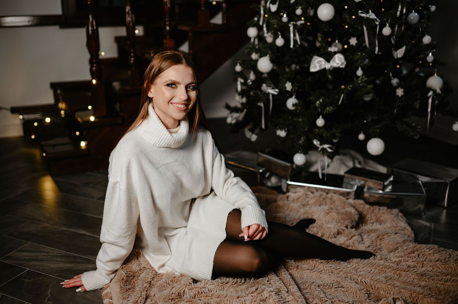 Smiling woman in white sweater sitting on fur rug by decorated Christmas tree.