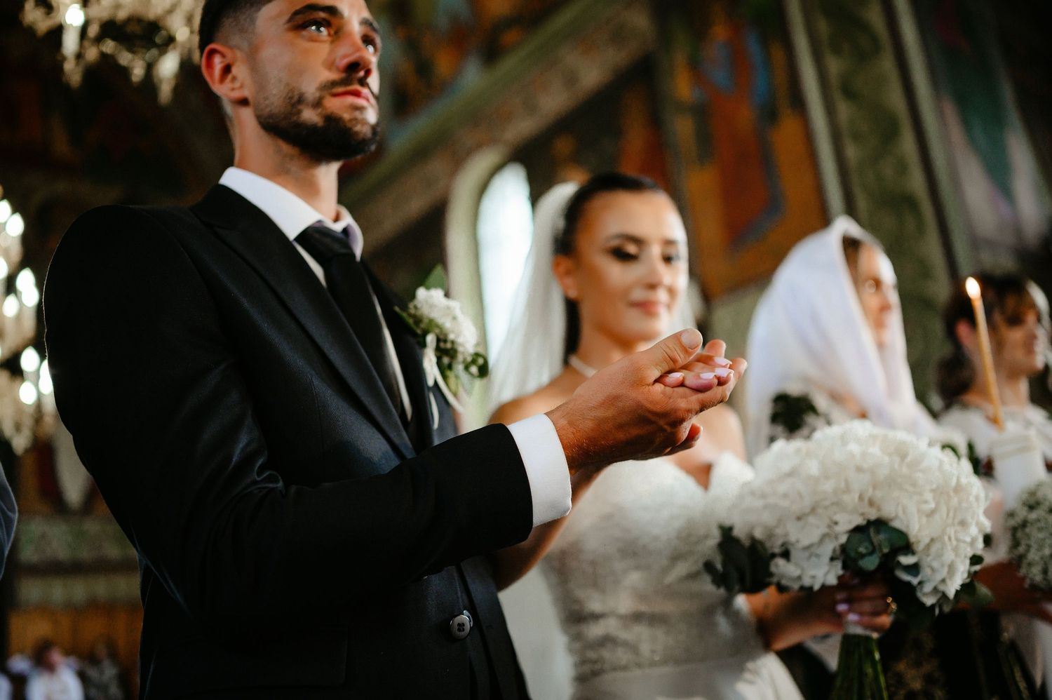 Groom in black suit holds wedding ring beside veiled bride with white bouquet in ornate church.
