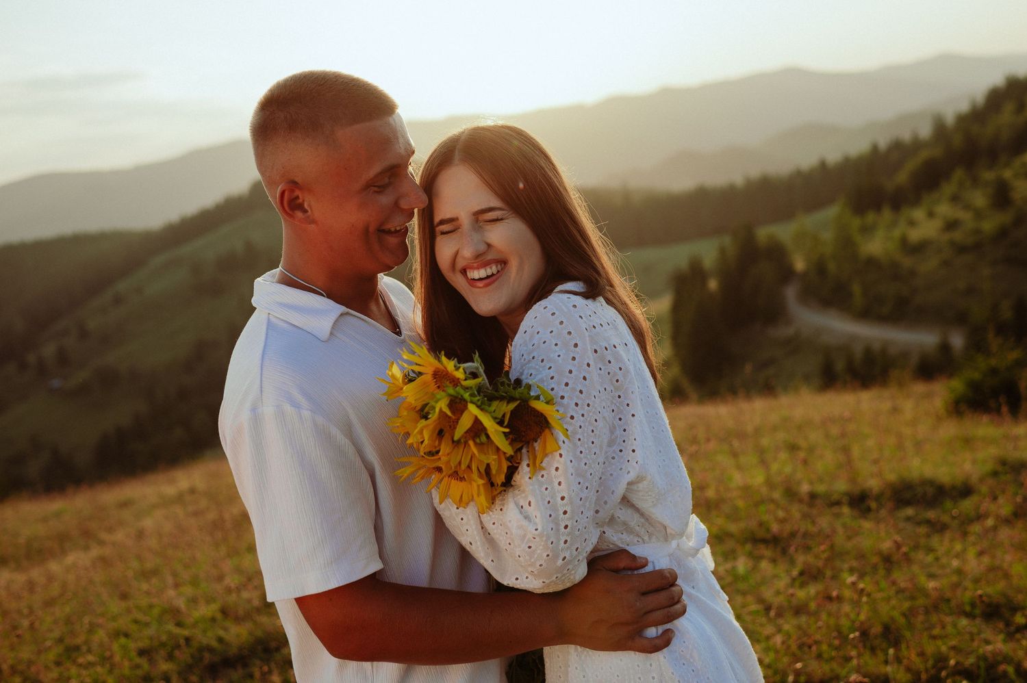 Happy couple embracing and laughing at sunset in scenic mountain meadow with sunflowers.