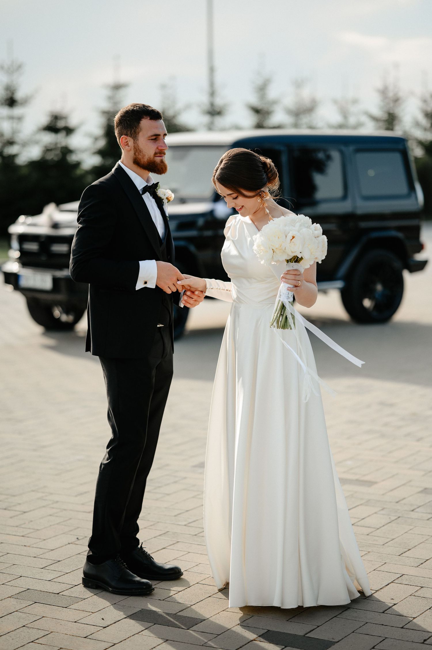 Bride and groom holding hands outdoors near black Mercedes G-Wagon on wedding day.
