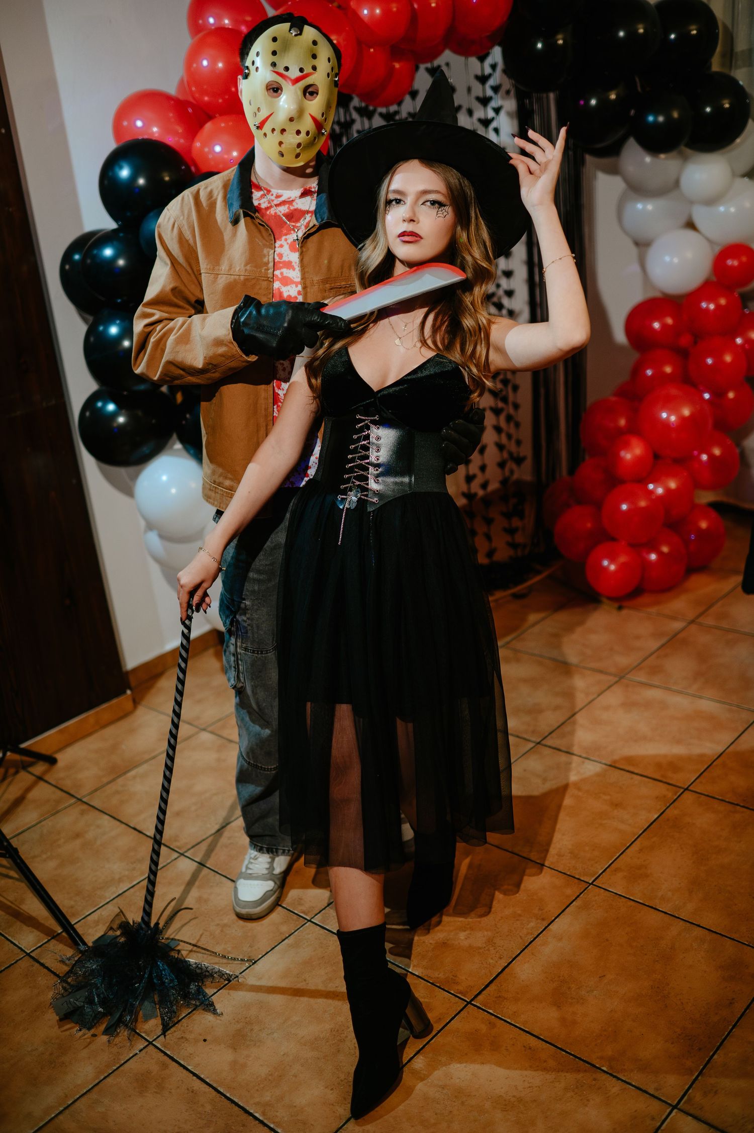 A couple in Halloween costumes, Jason Voorhees and a witch, pose at a party with red and black balloons.