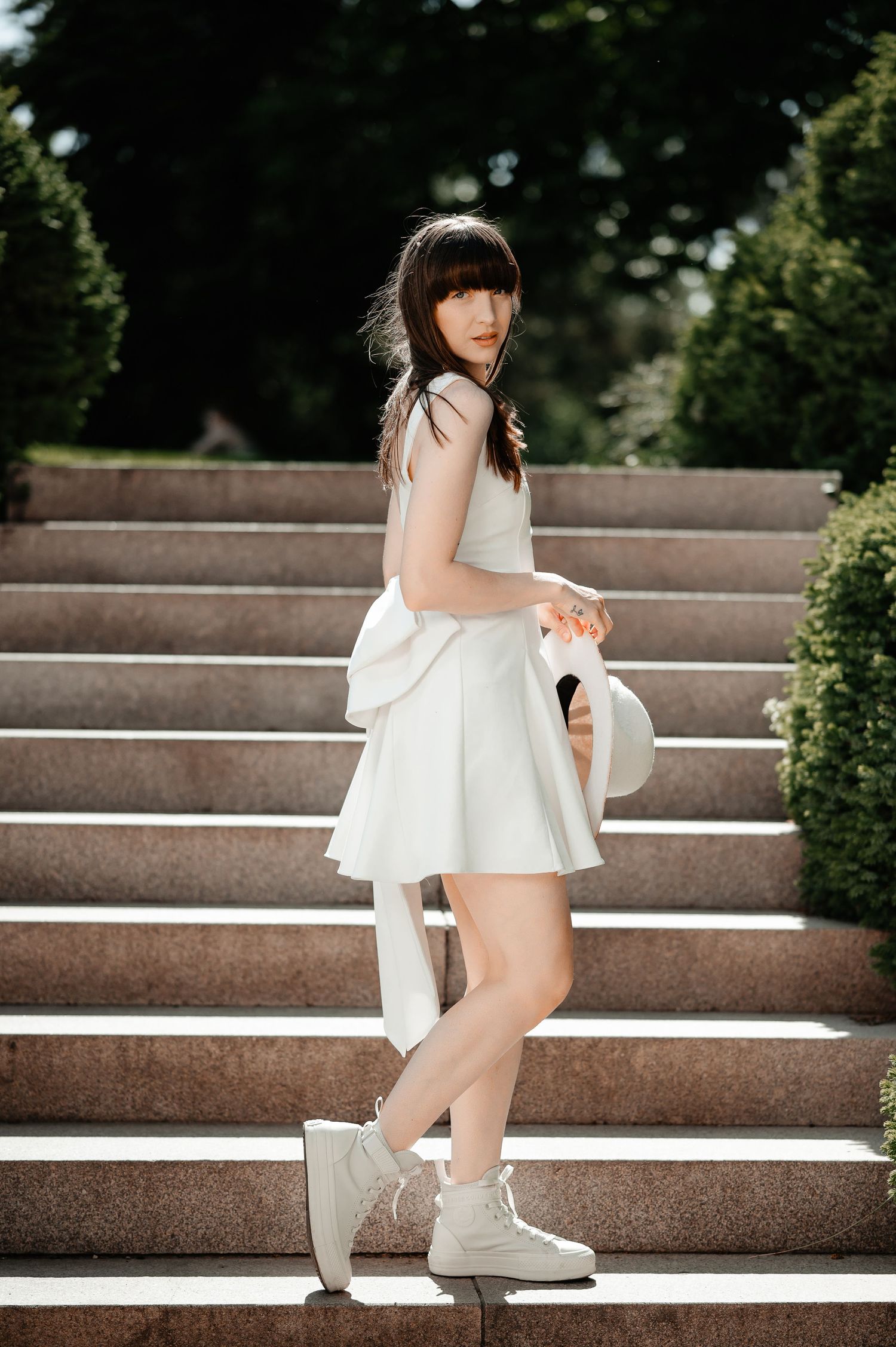 Stylish woman in white dress and sneakers posing on marble steps with hat in hand.