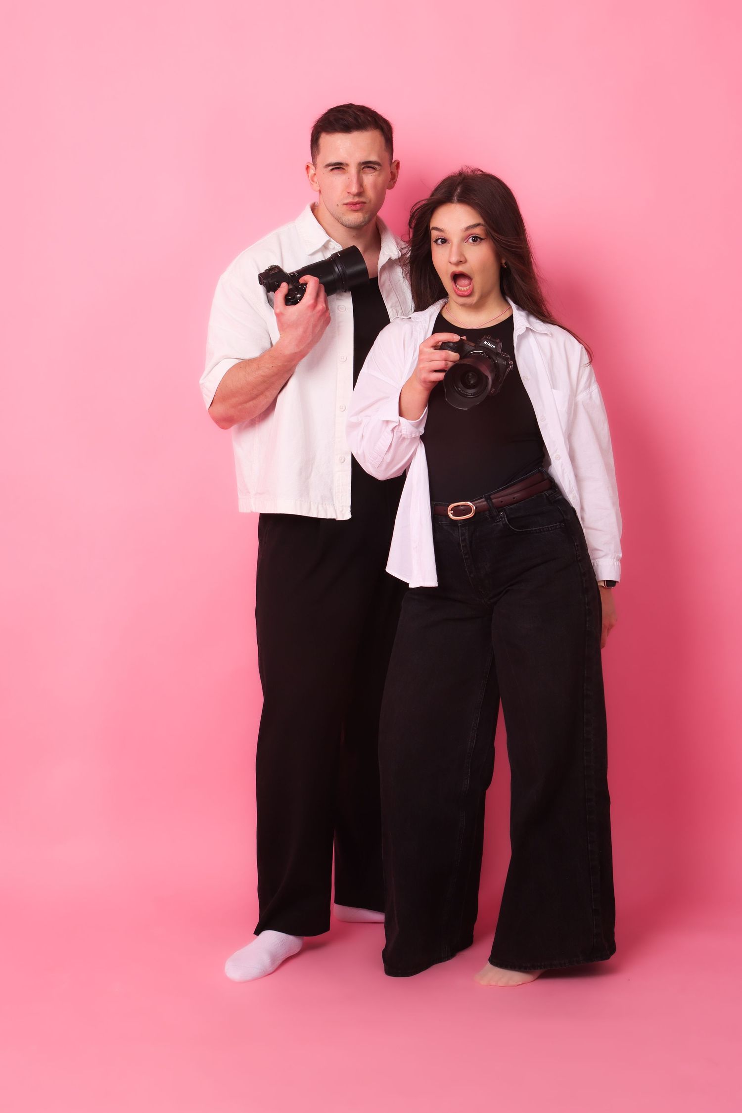 A man and woman in matching black and white outfits hold cameras against a pink background.