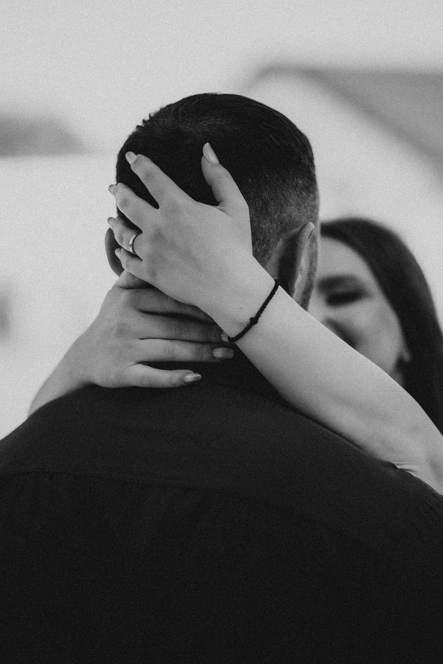 Close-up black and white photo of a woman's hand with ring embracing a man's neck.