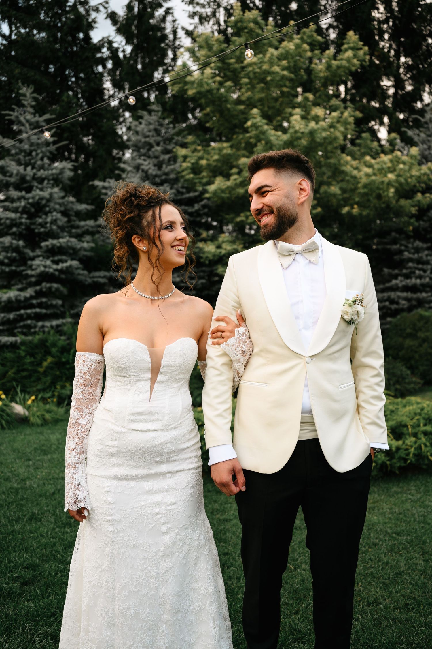 A smiling bride in a white lace off-shoulder gown and groom in ivory tuxedo hold hands outdoors.