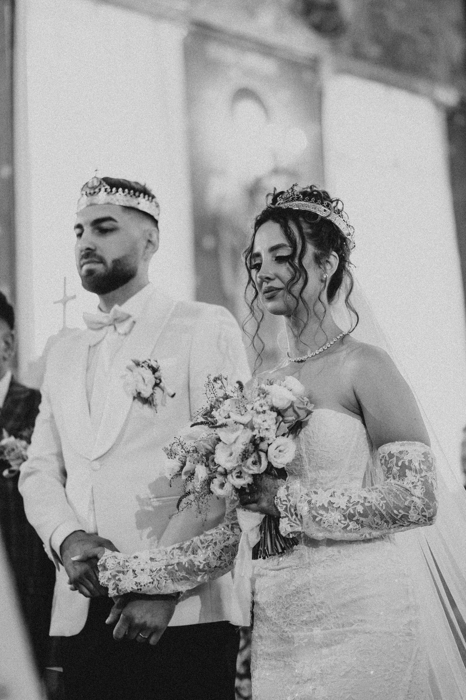 Black and white photo of a bride and groom wearing crowns at their wedding ceremony, bride holding a bouquet.