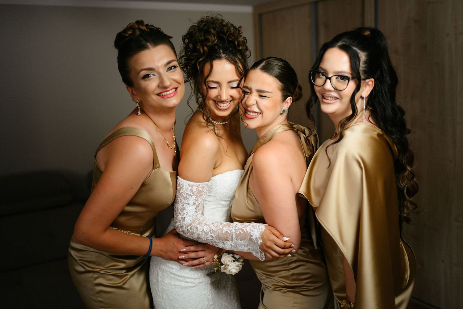 Bride in white lace gown embraced by three bridesmaids in gold satin dresses, smiling together before the wedding.
