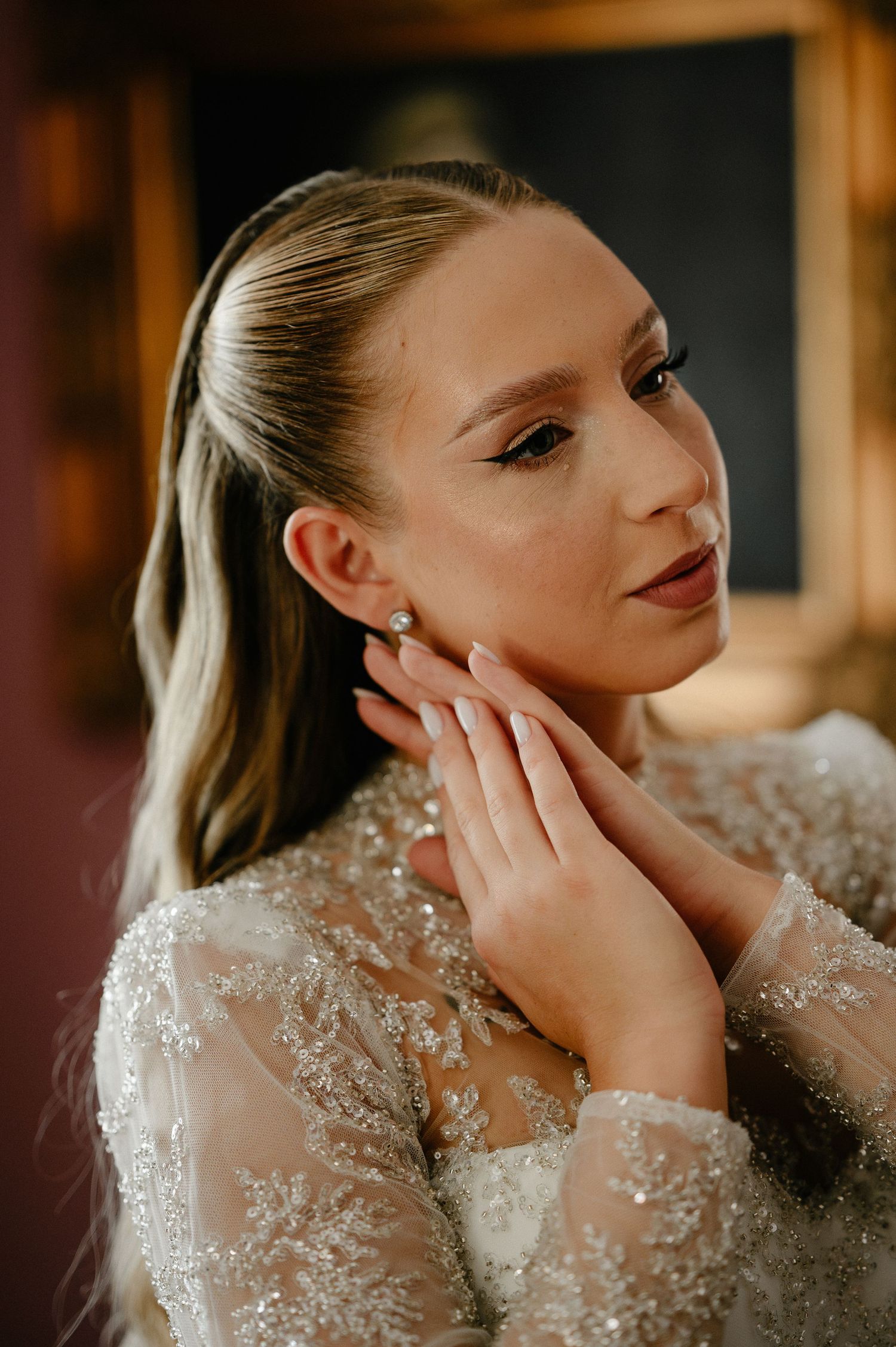 Elegant bride in beaded lace gown touching diamond earring with manicured hands in soft warm light.