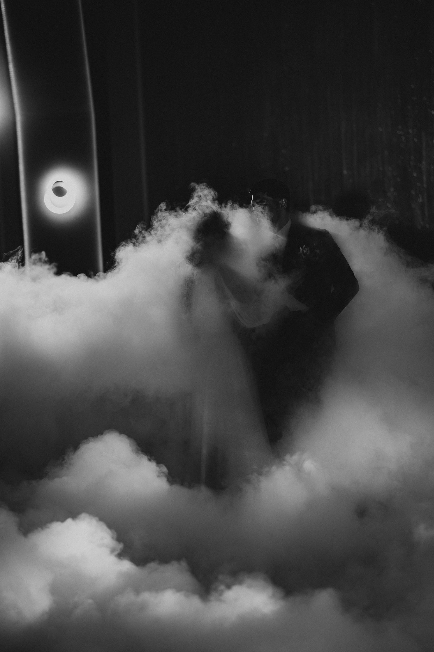 Dramatic black and white photo of bride and groom dancing surrounded by dry ice fog clouds.