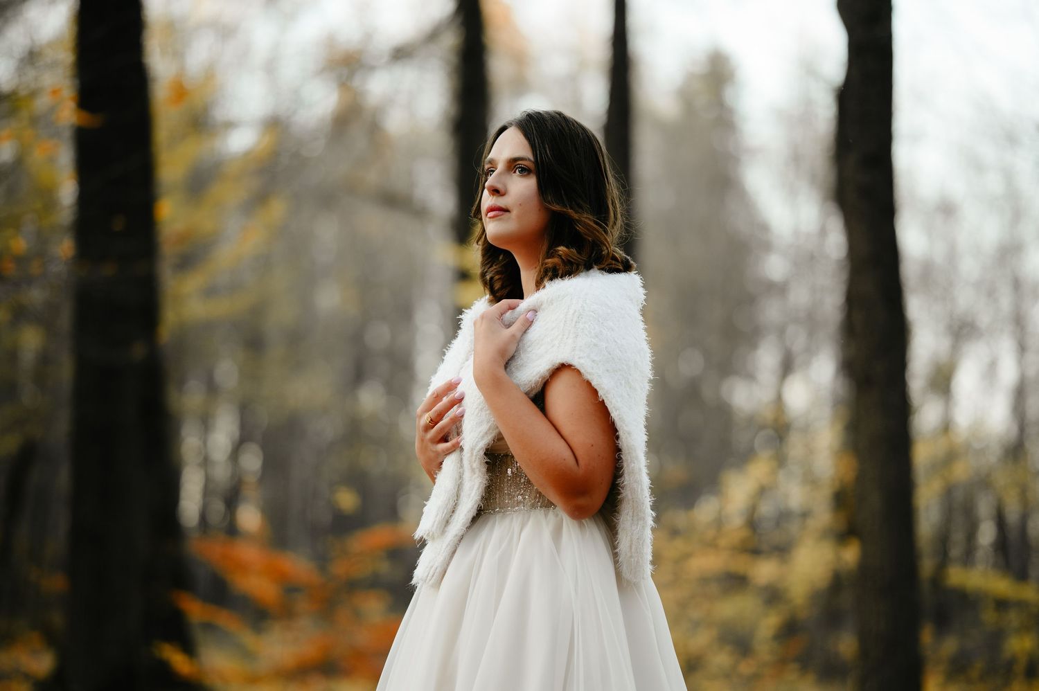 Bride in white gown and fur wrap poses dreamily in autumn forest with golden foliage backdrop.