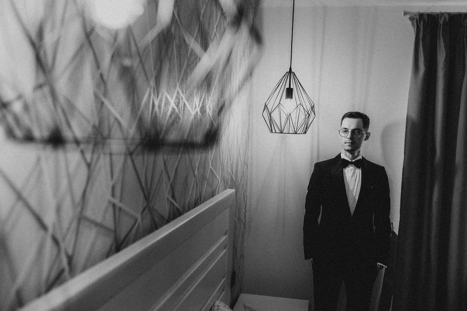 A groom in a black tuxedo and bow tie stands thoughtfully in a room with a geometric pendant light.