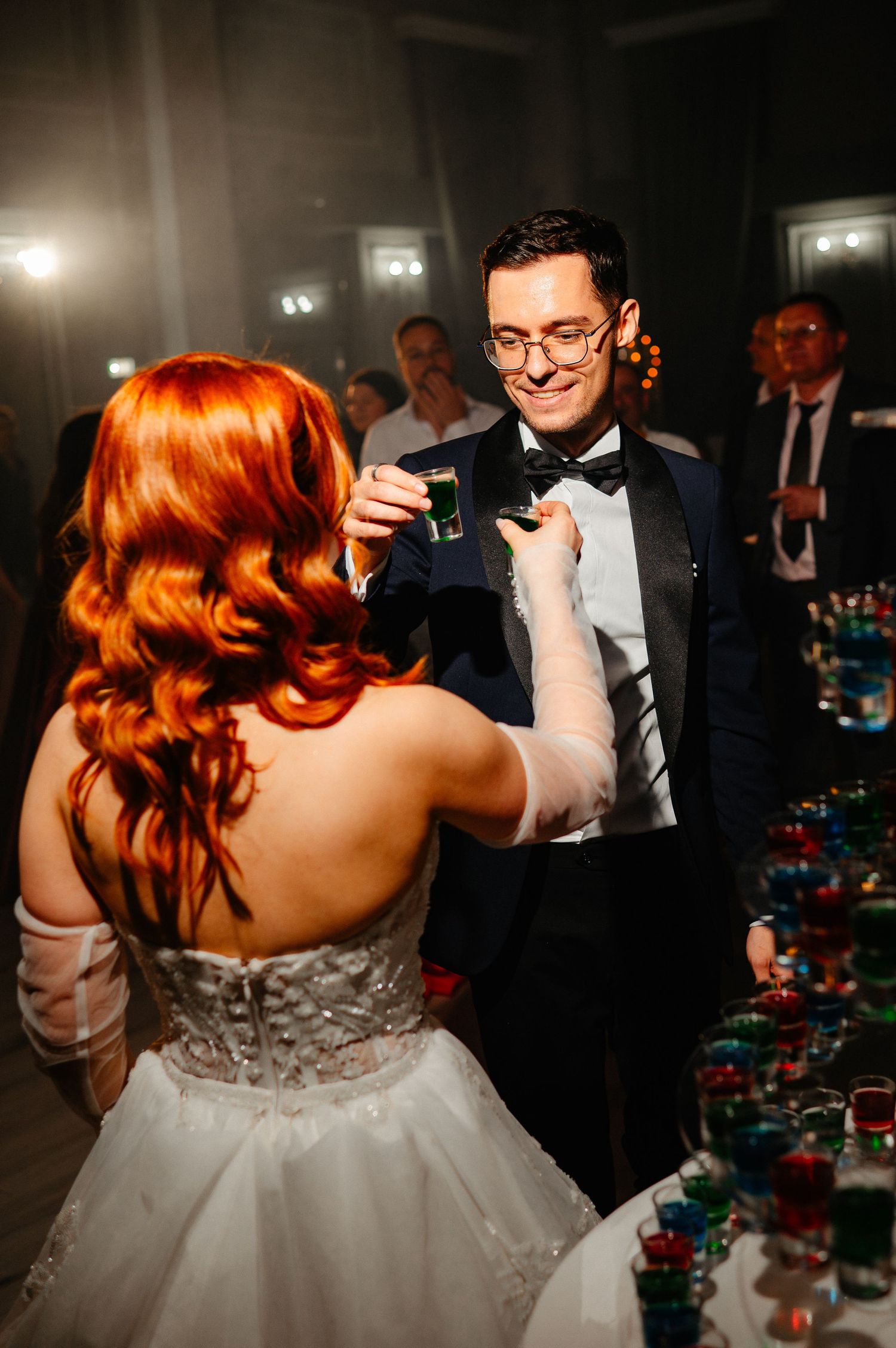 A bride with red hair in a white gown toasts with a groom in a tuxedo at their wedding reception.