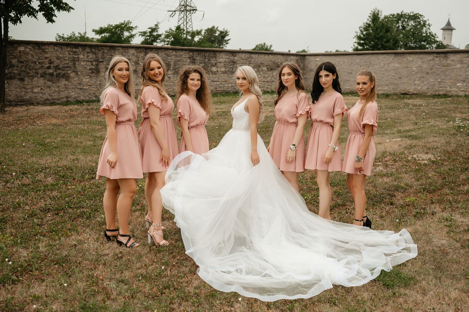 Bride in a white gown poses with six bridesmaids wearing matching dusty pink dresses outdoors.