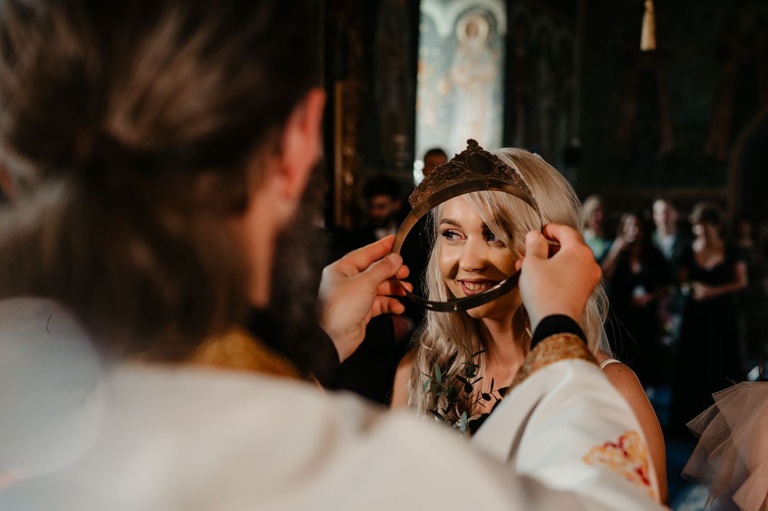 Orthodox priest placing a ceremonial crown on a smiling blonde bride during wedding ceremony.