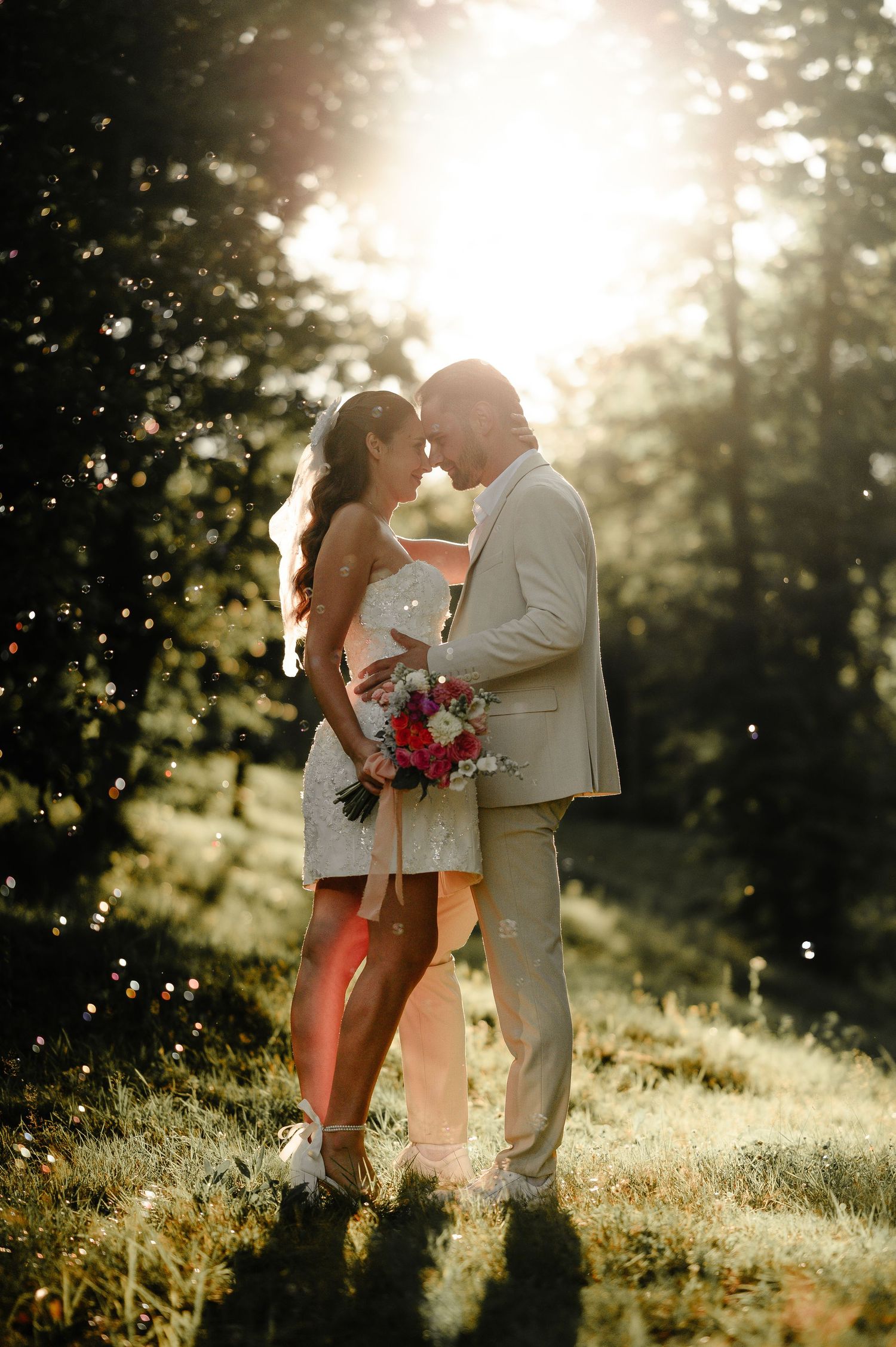 Bride and groom sharing intimate moment in golden sunlight surrounded by lush green trees on wedding day.