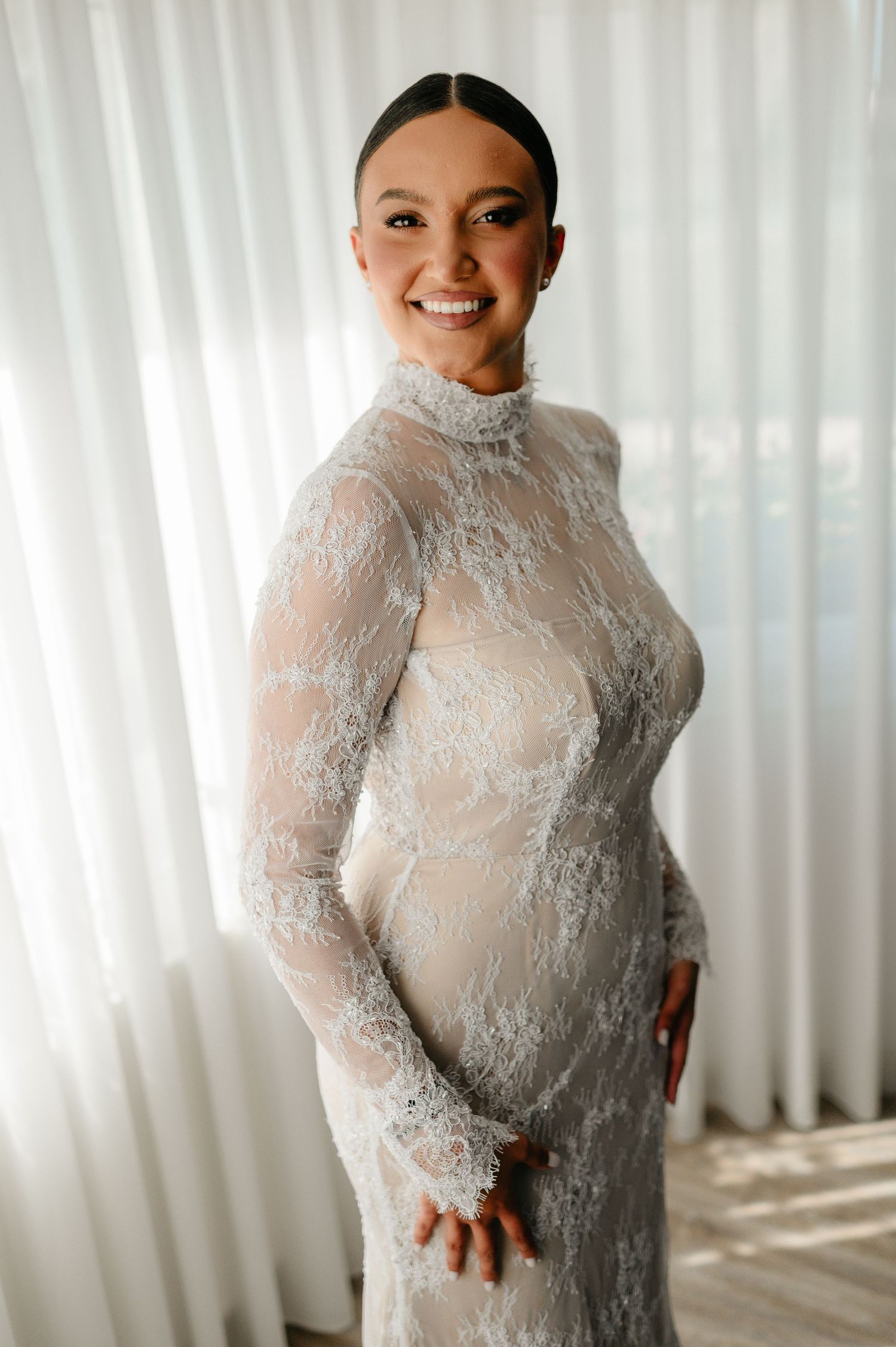 Smiling bride wearing a fitted high-neck long-sleeve lace wedding gown standing before sheer white curtains.