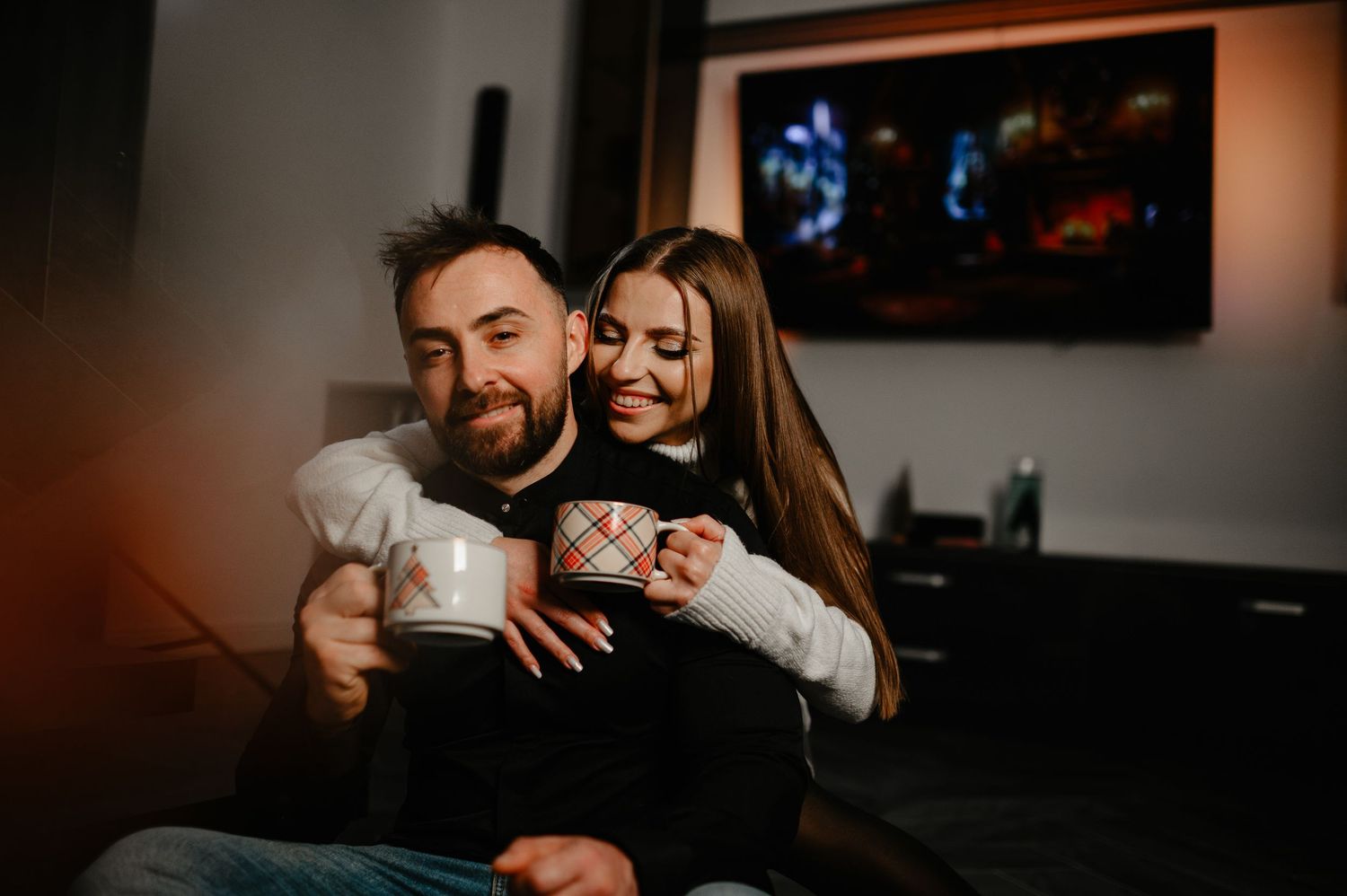 Couple cuddling on a couch holding mugs with a TV in the background.