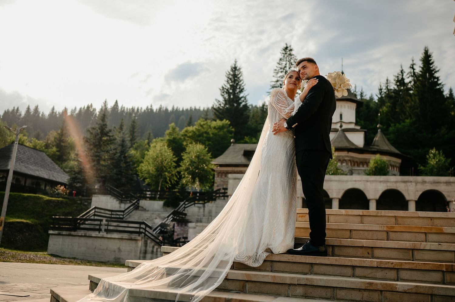 Bride and groom share a romantic embrace on stone steps with a church and forest backdrop on their wedding day.