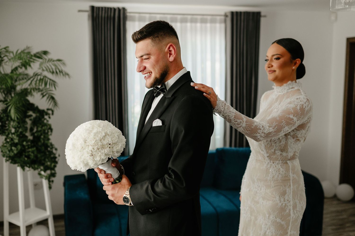 Bride in lace gown adjusting grooms black tuxedo bow tie before wedding ceremony.