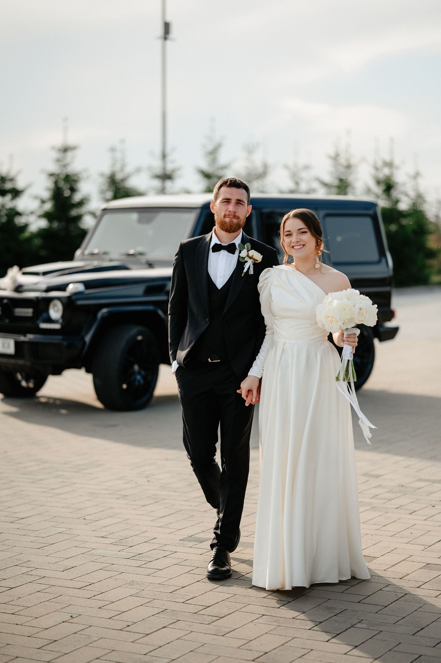 Bride and groom walking hand in hand outdoors near black Mercedes G-Wagon.