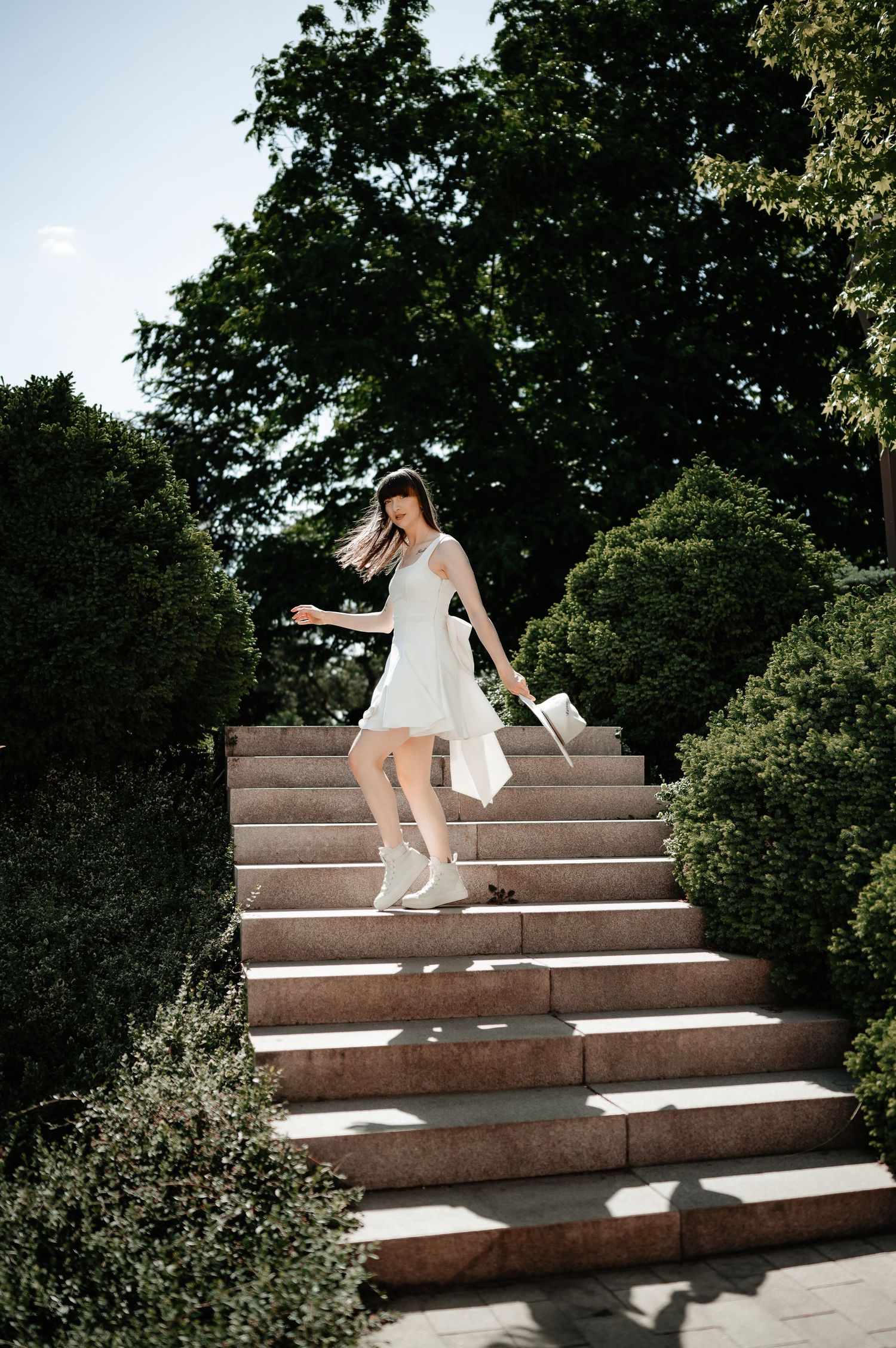 Young woman in white dress and sneakers descending stone stairs in a lush green park.