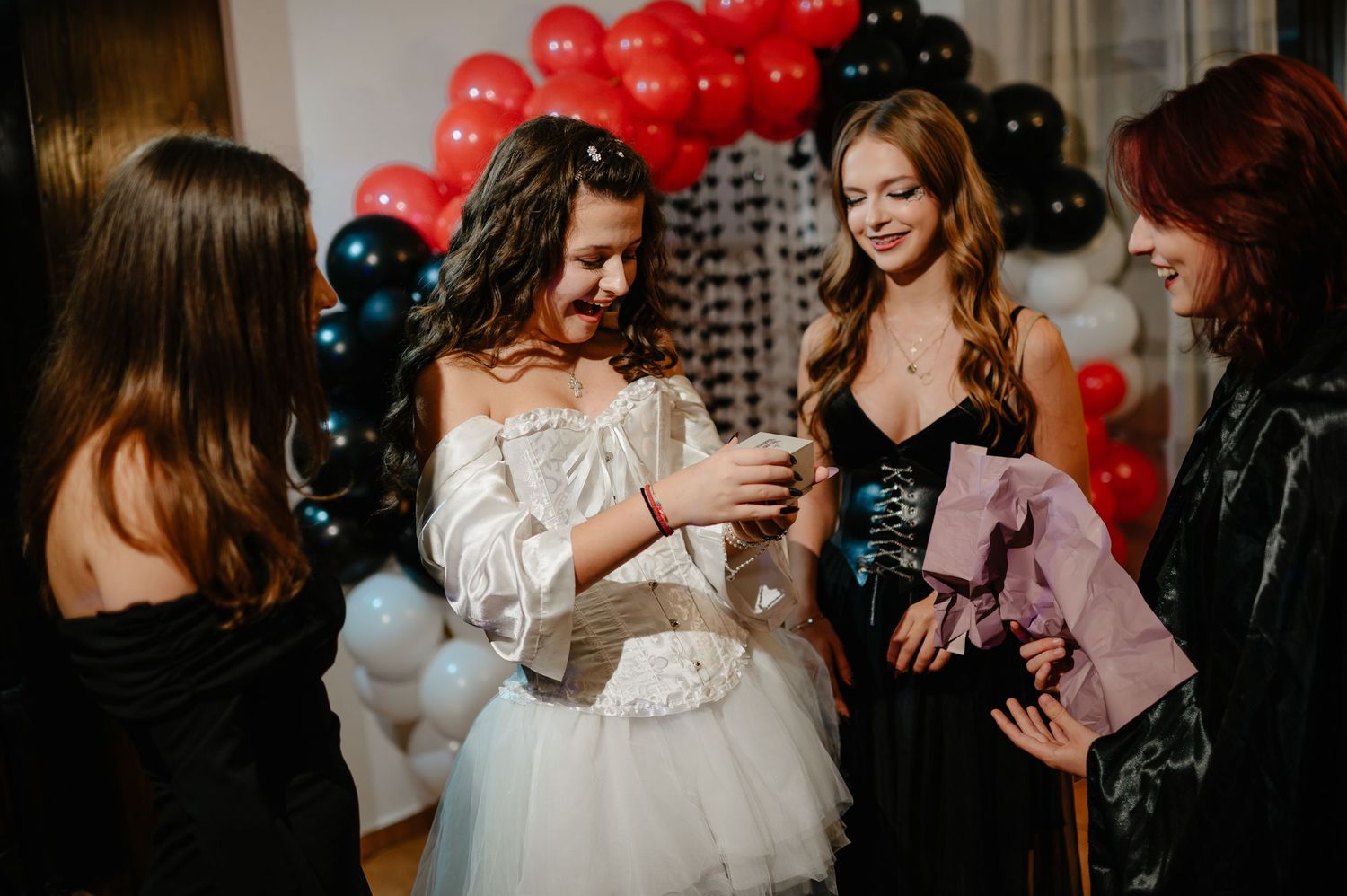 Group of women in Halloween costumes laughing and socializing near red and black balloon arch.