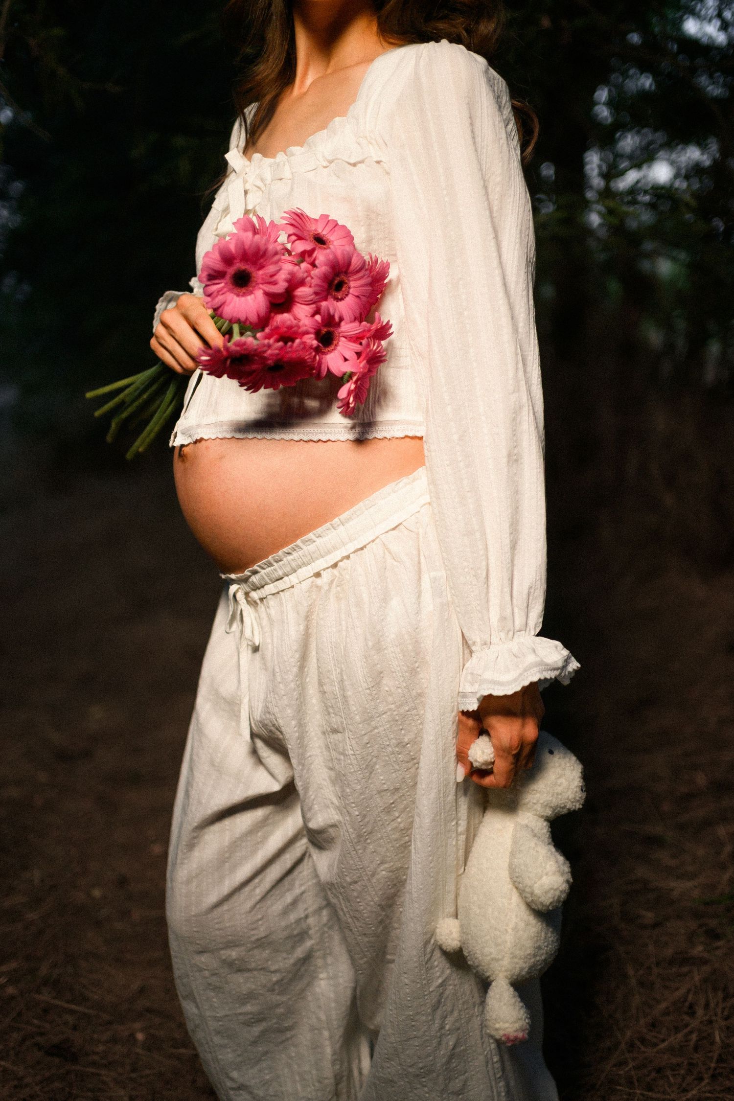 Pregnant woman in white linen outfit holding pink gerbera bouquet and teddy bear outdoors.