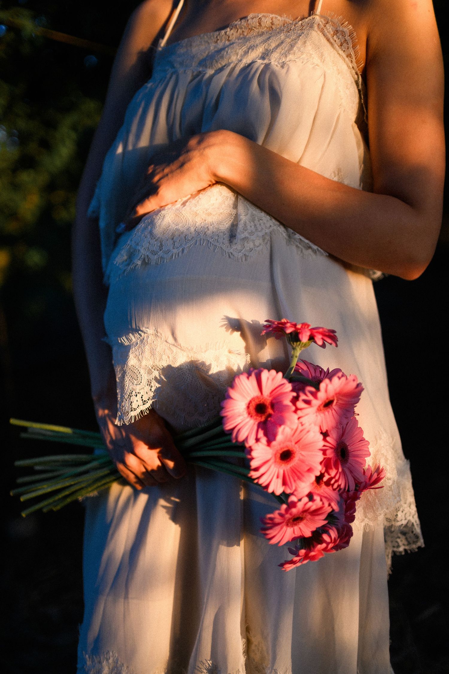Pregnant woman in white lace dress holding pink gerbera daisy bouquet in golden sunset light.
