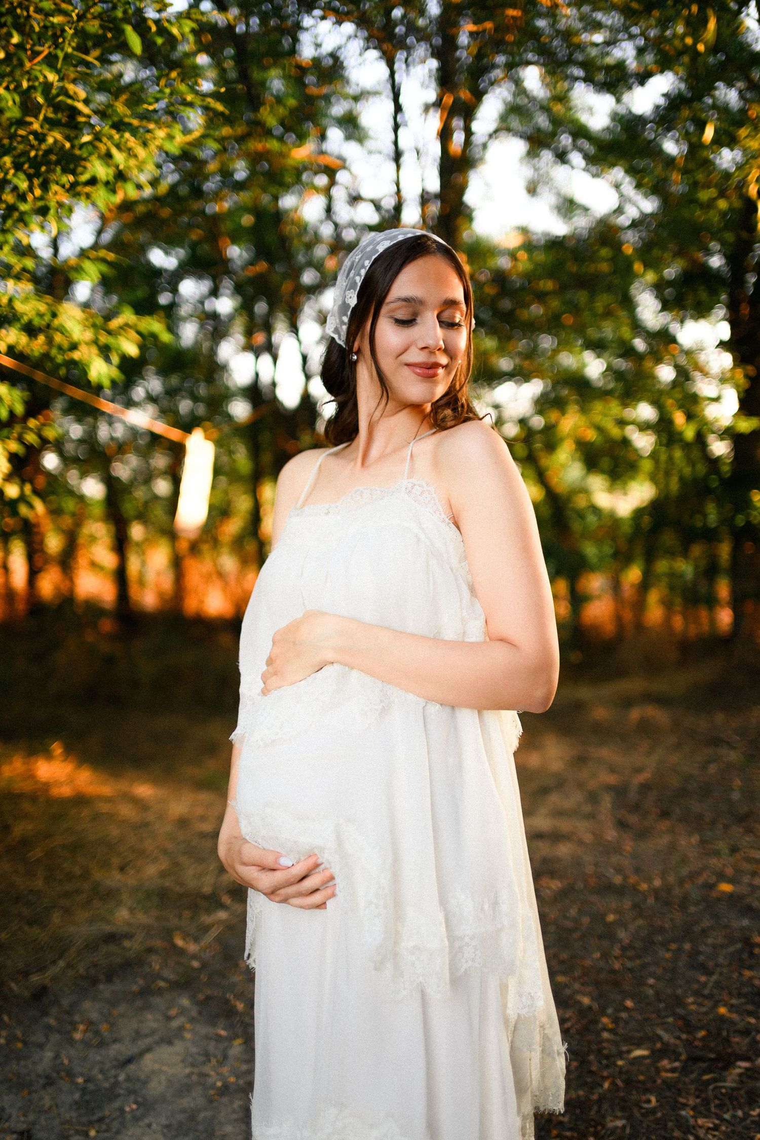 Pregnant woman in white lace dress and headpiece cradling her belly outdoors in golden sunlight among green trees.