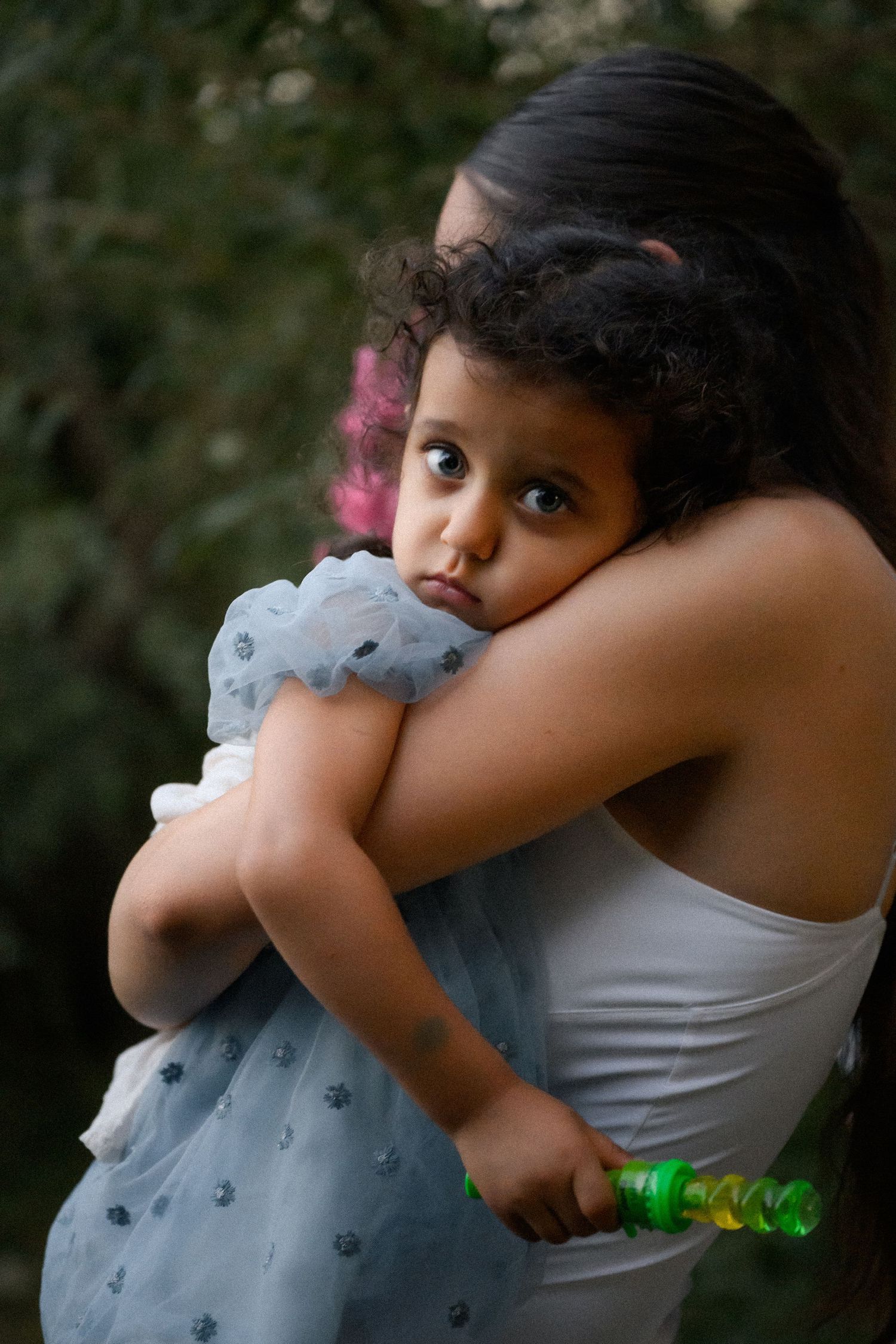 A woman in a white top holds a young girl in a blue dress outdoors, the child gazing at the camera.