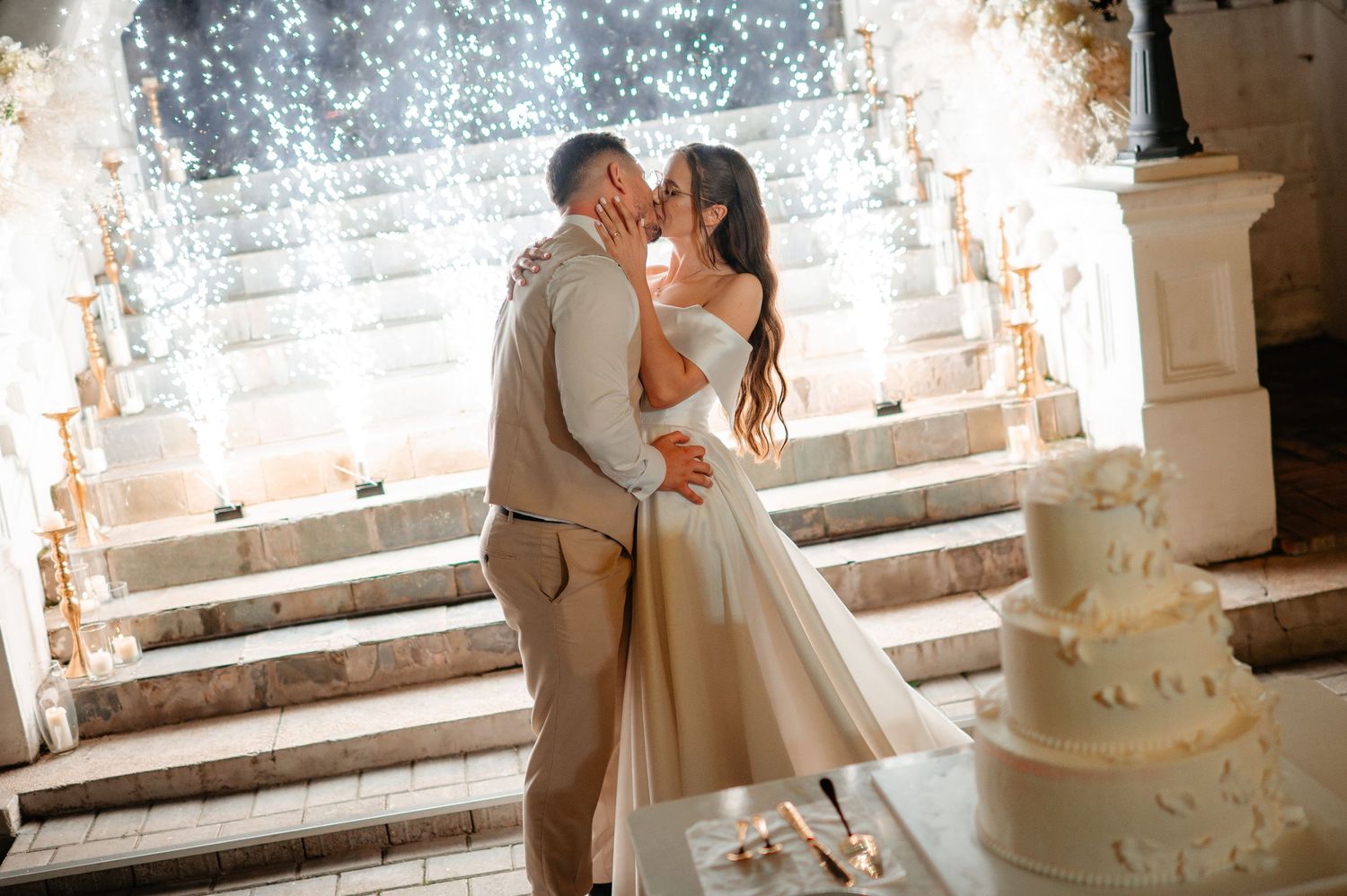 Bride and groom share romantic kiss beside wedding cake with stunning sparkling light display on steps.
