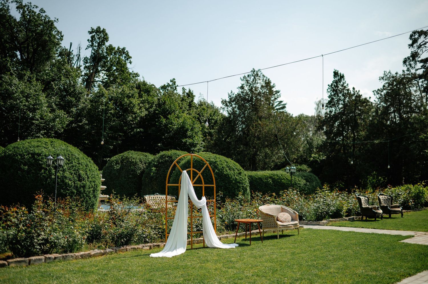 Outdoor wedding ceremony arch with white draping in a lush green garden setting.