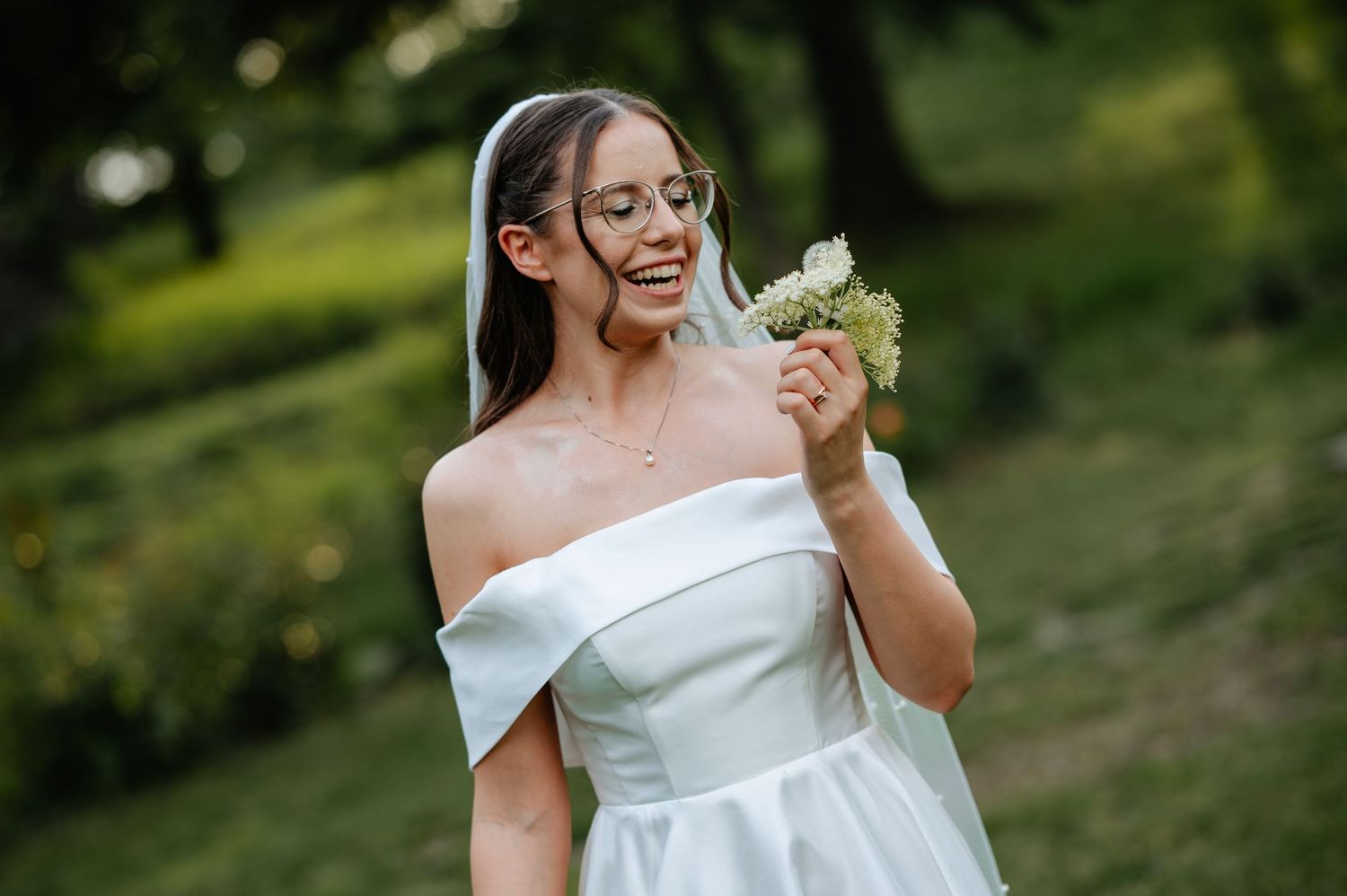 Smiling bride in off-shoulder white gown holding a small bouquet in a sunlit garden.