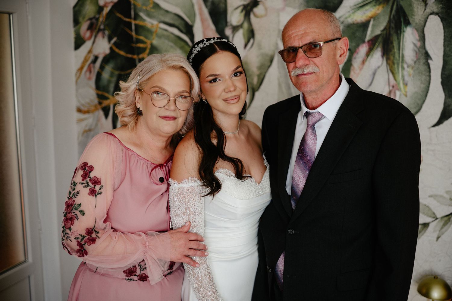 Happy bride in white lace gown posing with parents in front of tropical botanical wallpaper backdrop.