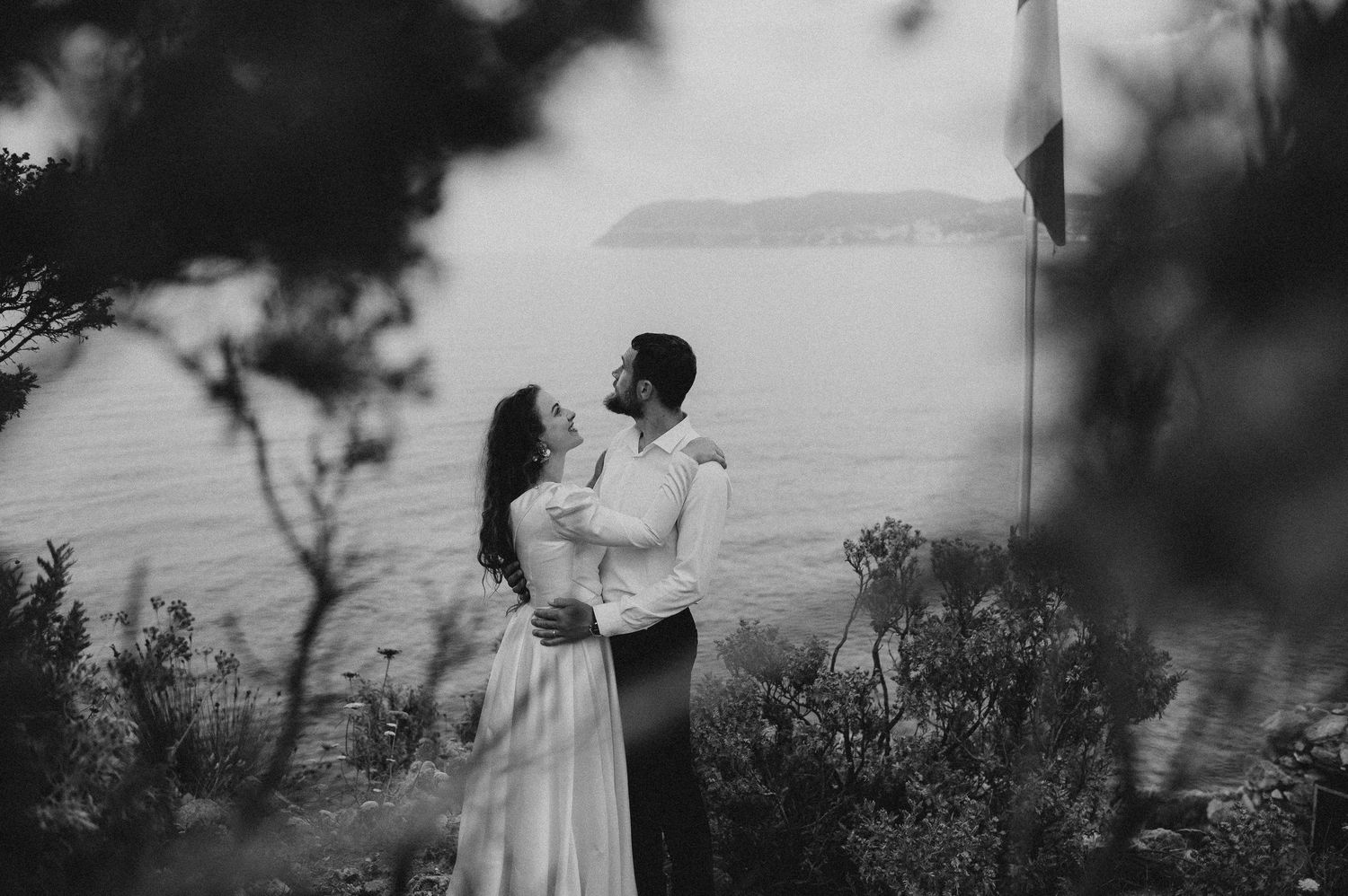 Black and white romantic photo of wedding couple embracing near coastal trees with sea behind them.