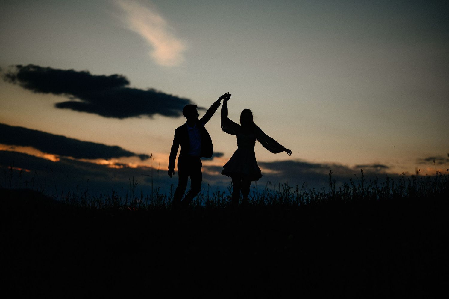 Silhouette of a couple dancing at sunset with dramatic sky and raised hands against the horizon.