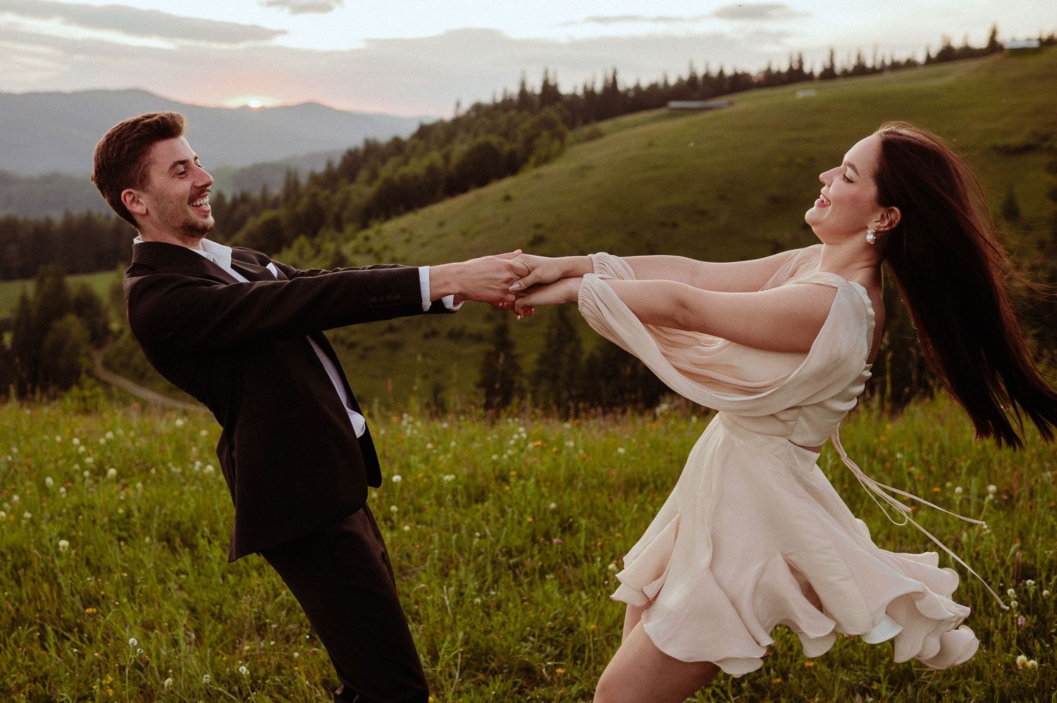 Joyful couple spinning together in a green meadow at sunset during engagement photo session.