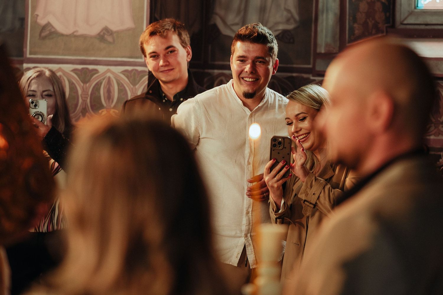 Guests holding candles and smiling during an Orthodox Christian baptism ceremony inside a church.