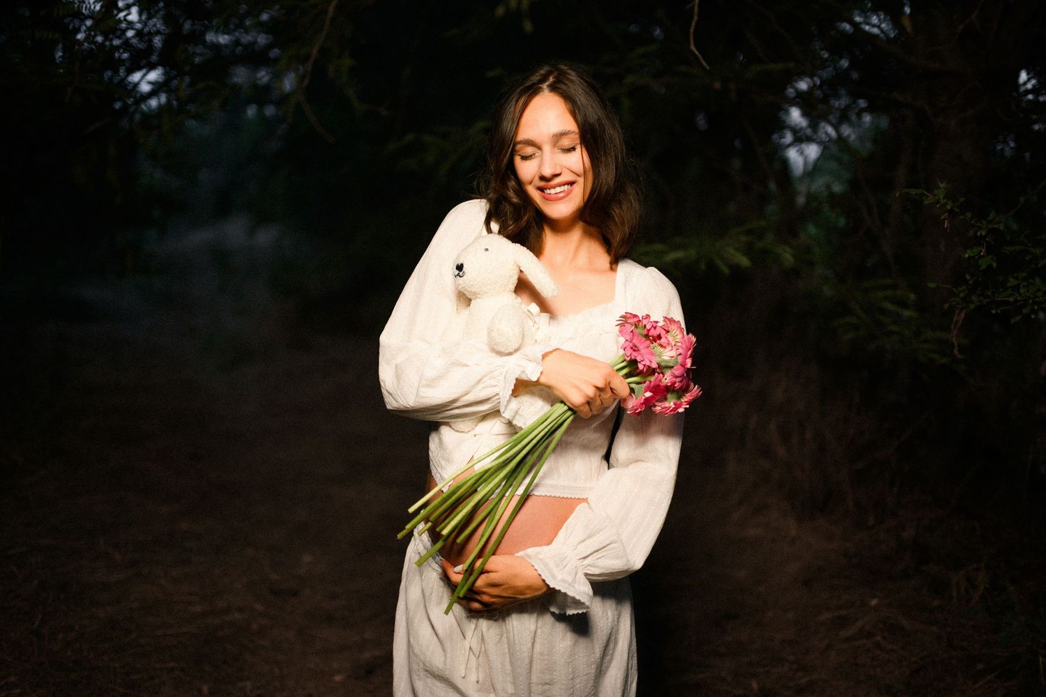 Smiling woman in white dress holding pink flowers and a white rabbit in a dark forest setting.