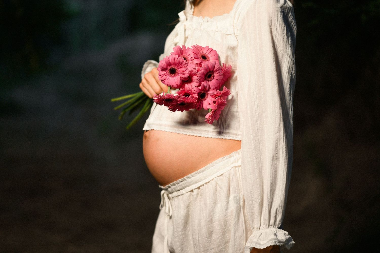 Pregnant woman in white dress holding pink gerbera daisy bouquet outdoors, showcasing baby bump in natural light.
