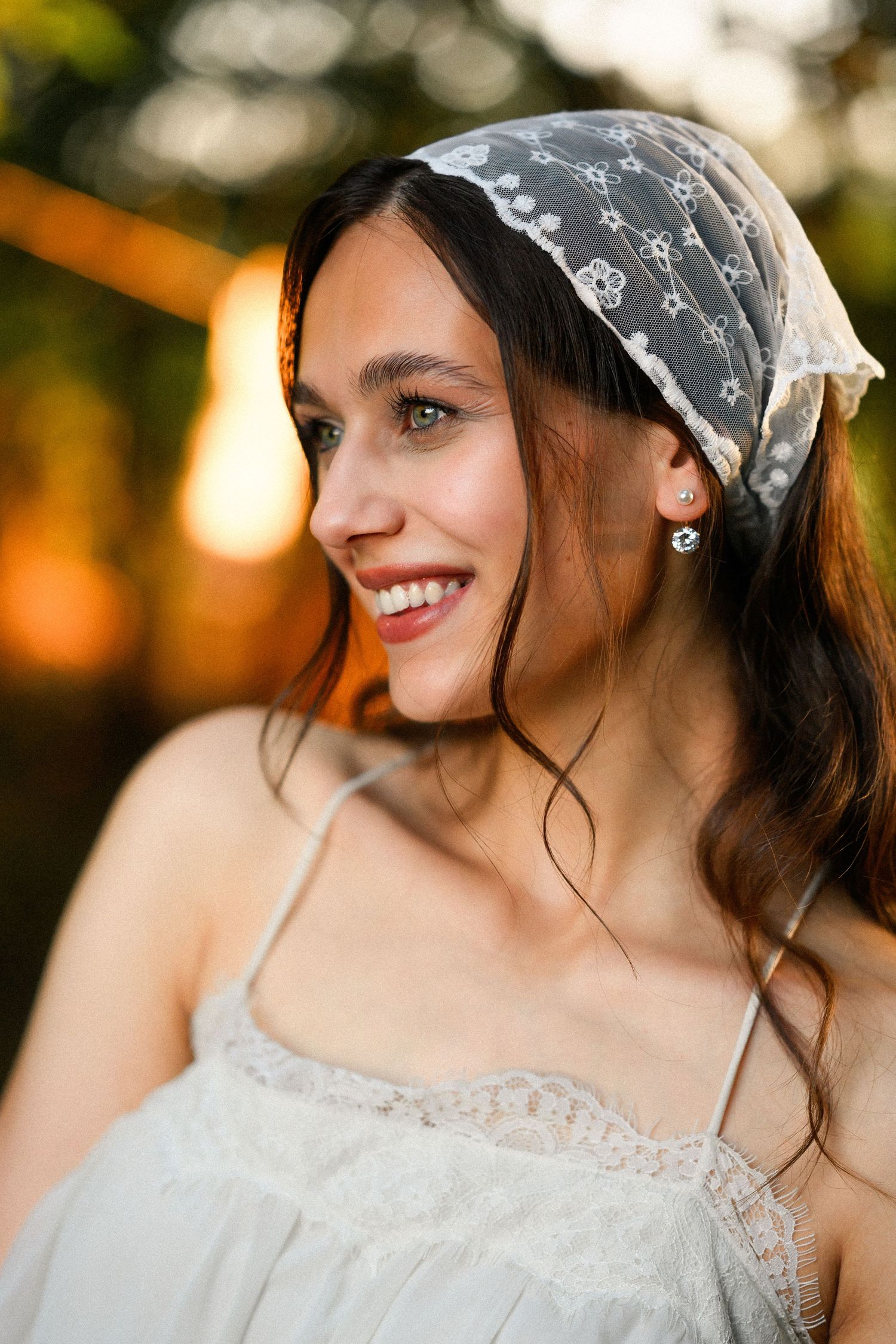 Smiling young woman wearing a white lace headscarf and floral earrings in a white dress, photographed outdoors at golden hour.