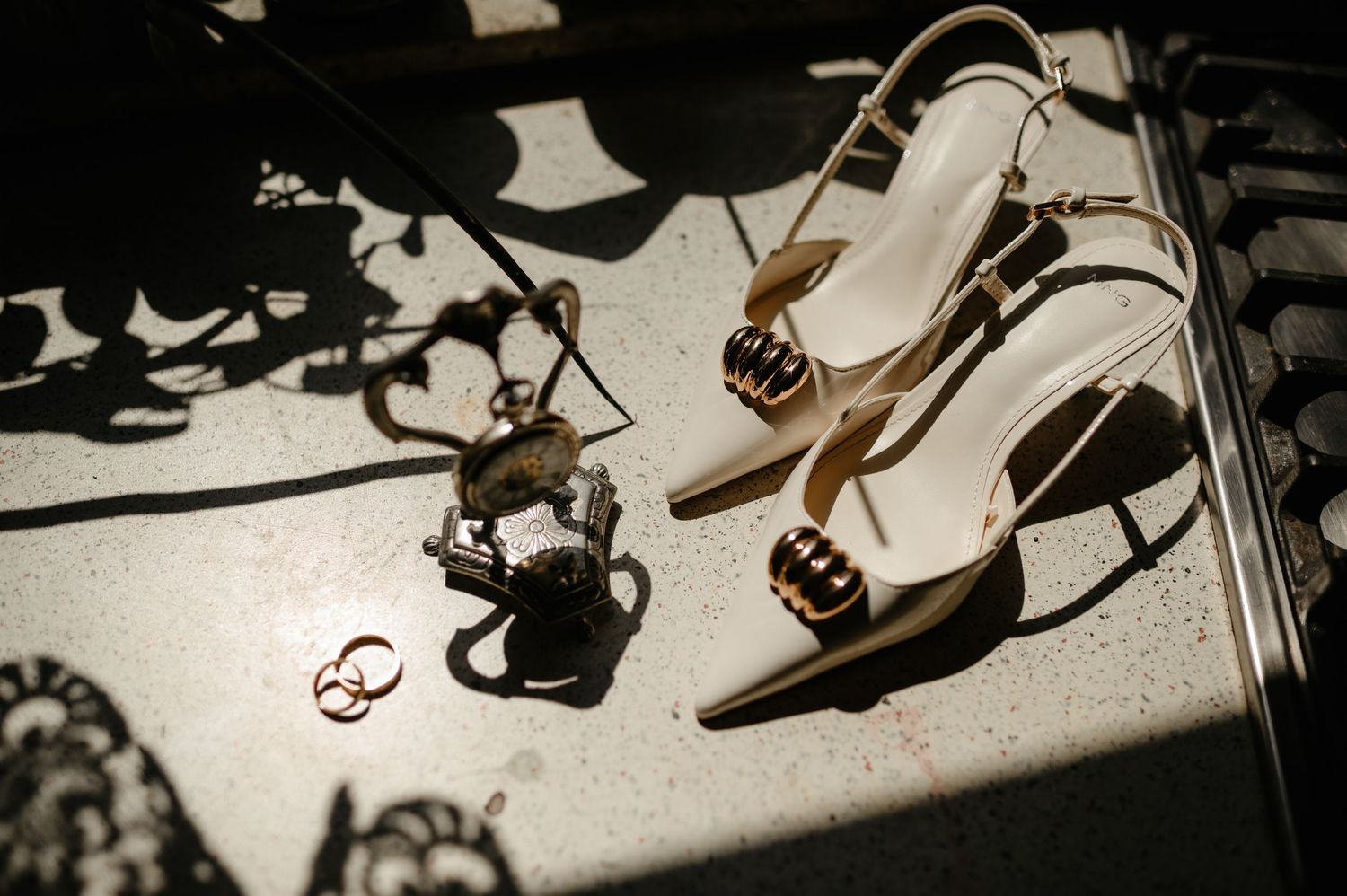 Elegant white bridal heels with gold accents beside wedding rings and a decorative figurine on a marble surface.