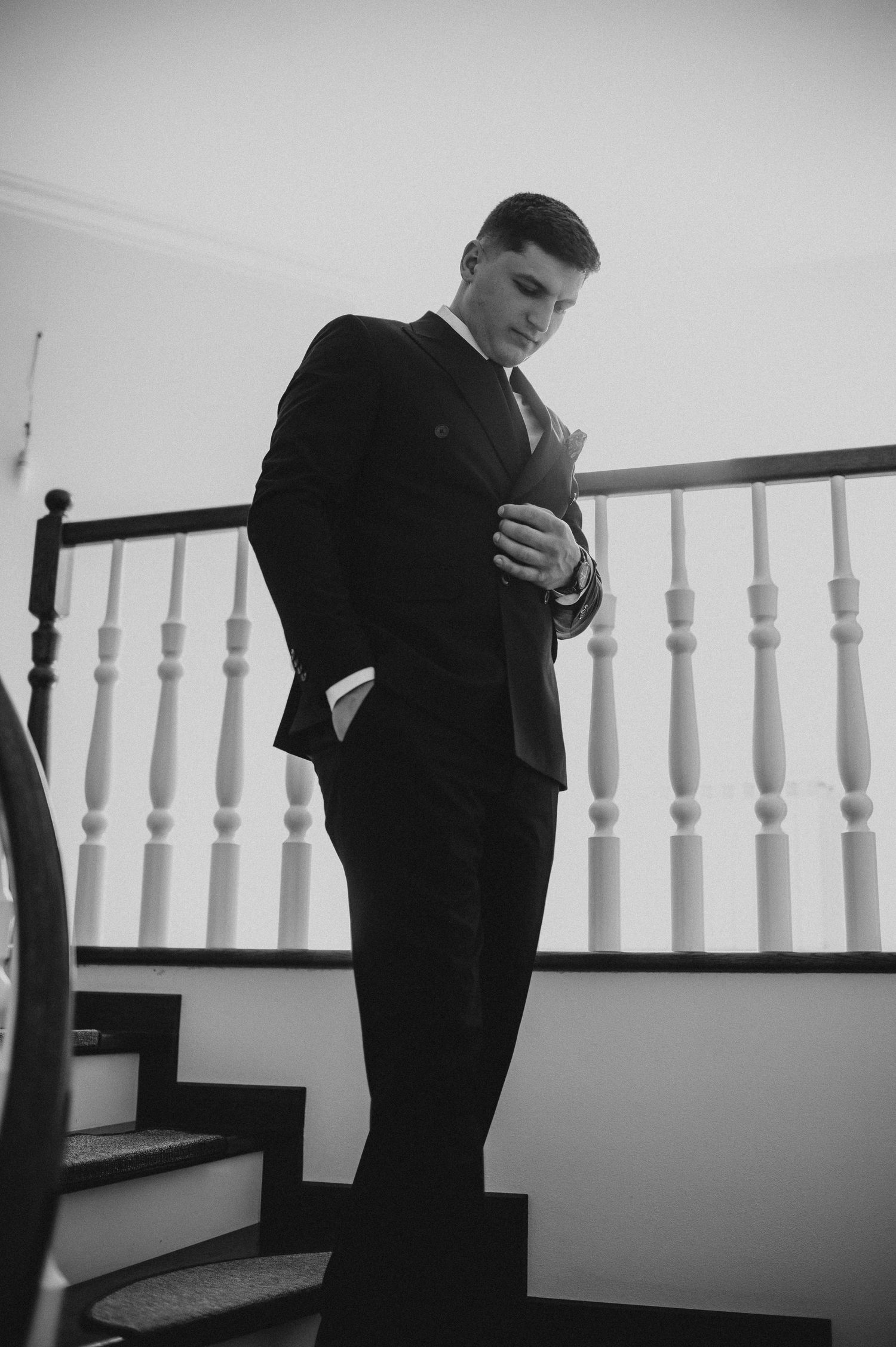 Groom in black suit adjusting jacket on staircase, black and white wedding photo.
