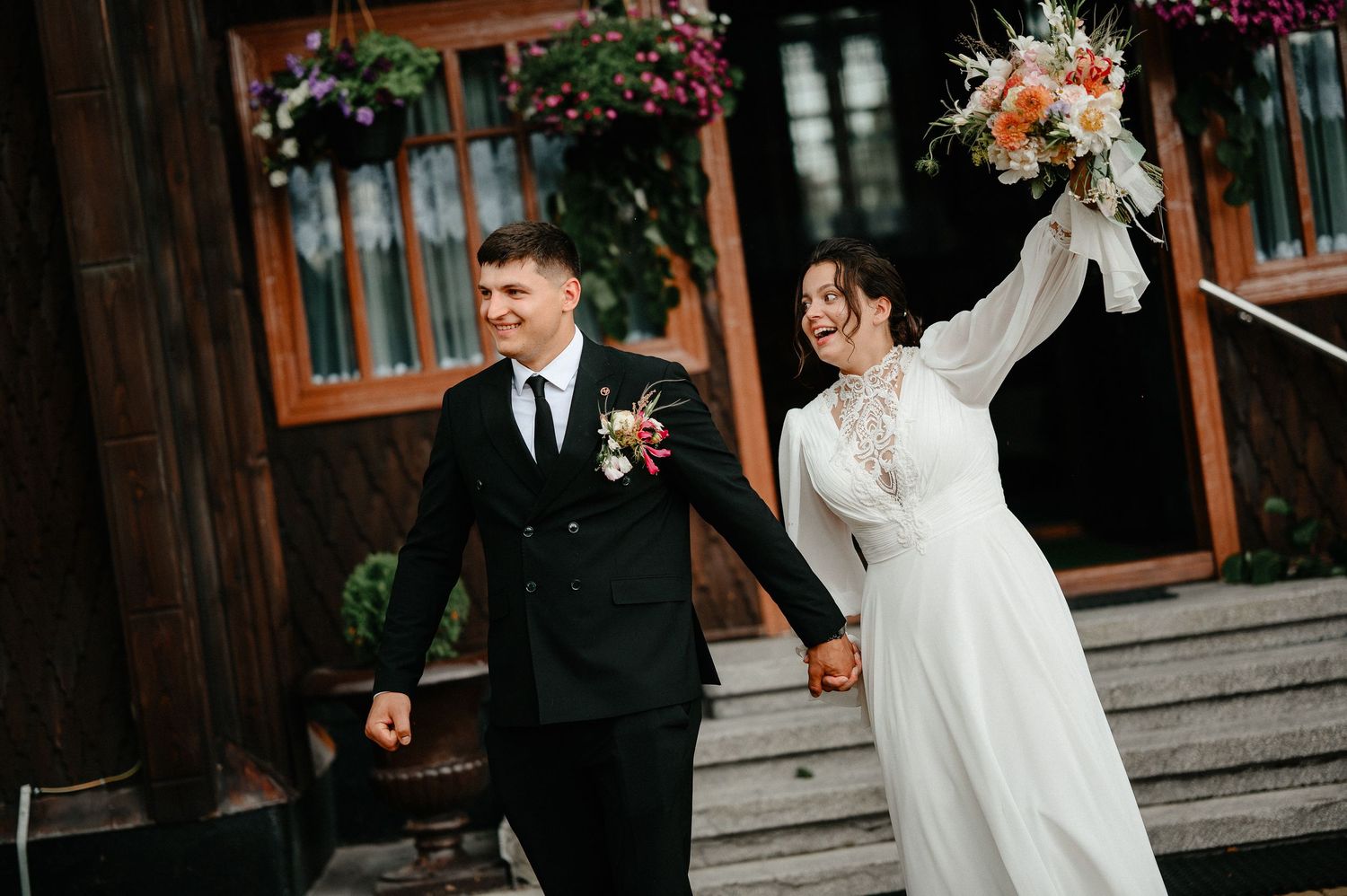 Happy newlyweds holding hands and celebrating outside a rustic wooden venue with colorful hanging flowers.