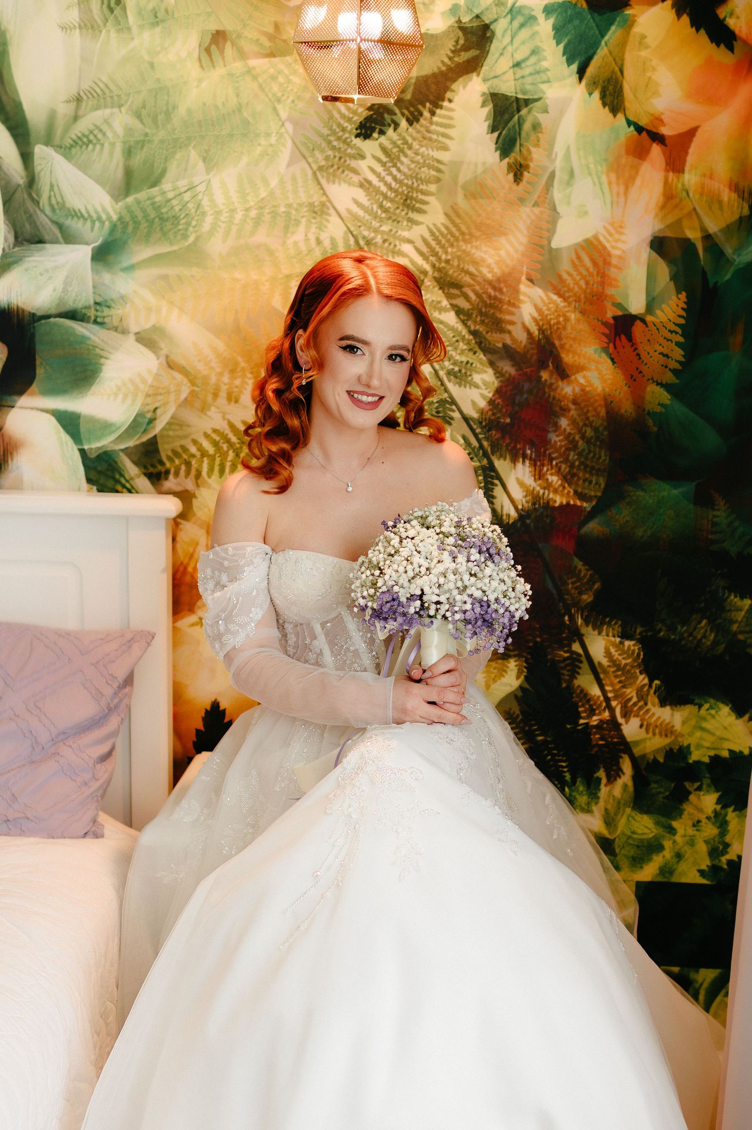 Smiling bride in white off-shoulder gown holding purple bouquet, seated before vibrant tropical floral wallpaper.