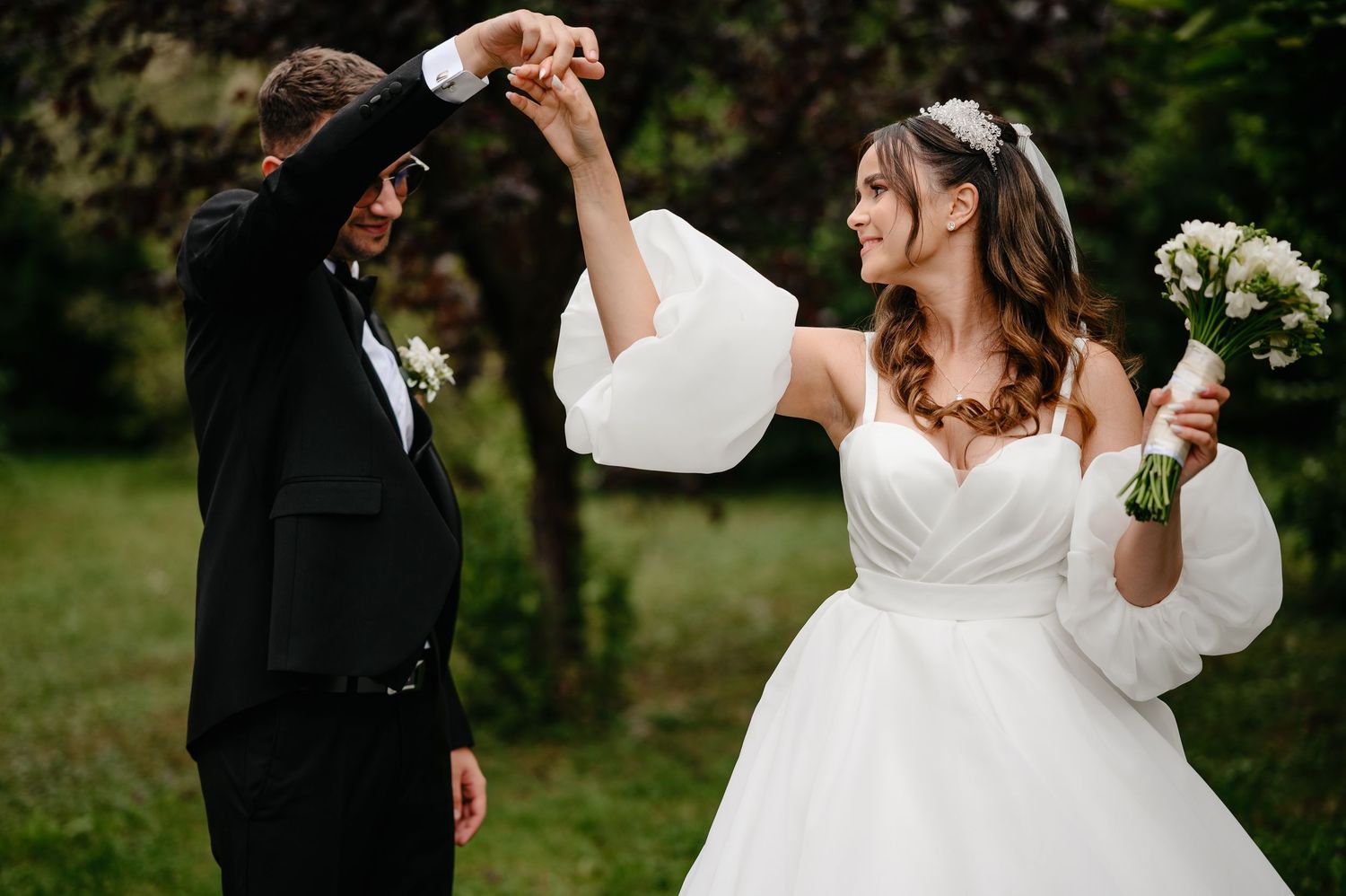 Joyful newlyweds dancing outdoors, bride in white off-shoulder gown holding bouquet, groom in black tuxedo.